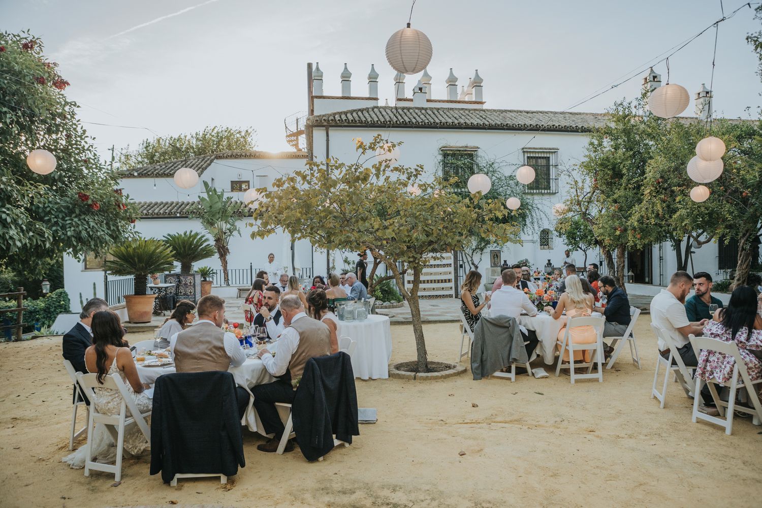 Guests enjoy an outdoor wedding reception with tables and paper lanterns at a rustic venue.