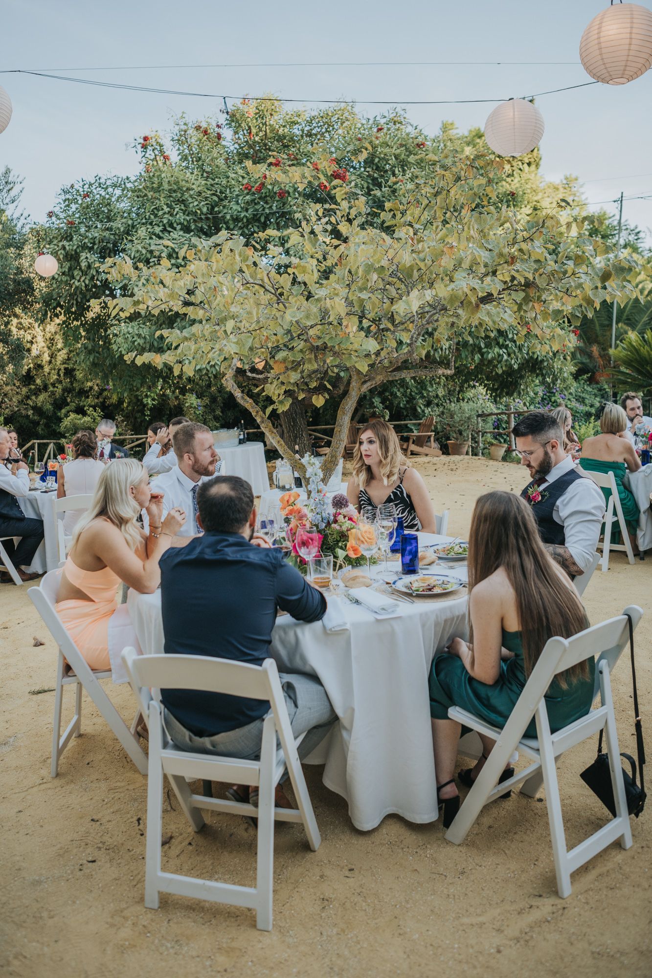 Outdoor wedding reception under a large tree with paper lanterns and white folding chairs.