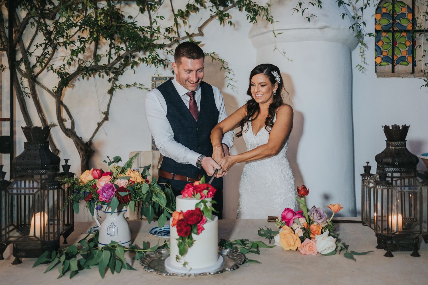 A bride and groom cut their floral wedding cake together outdoors against a white wall with lantern decorations.