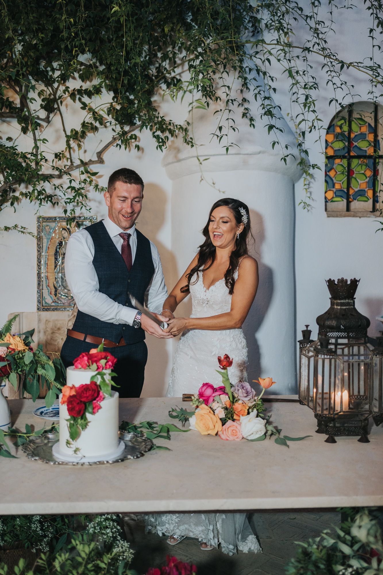 Newlyweds cut their elegant white wedding cake decorated with pink and coral roses at a romantic garden venue.
