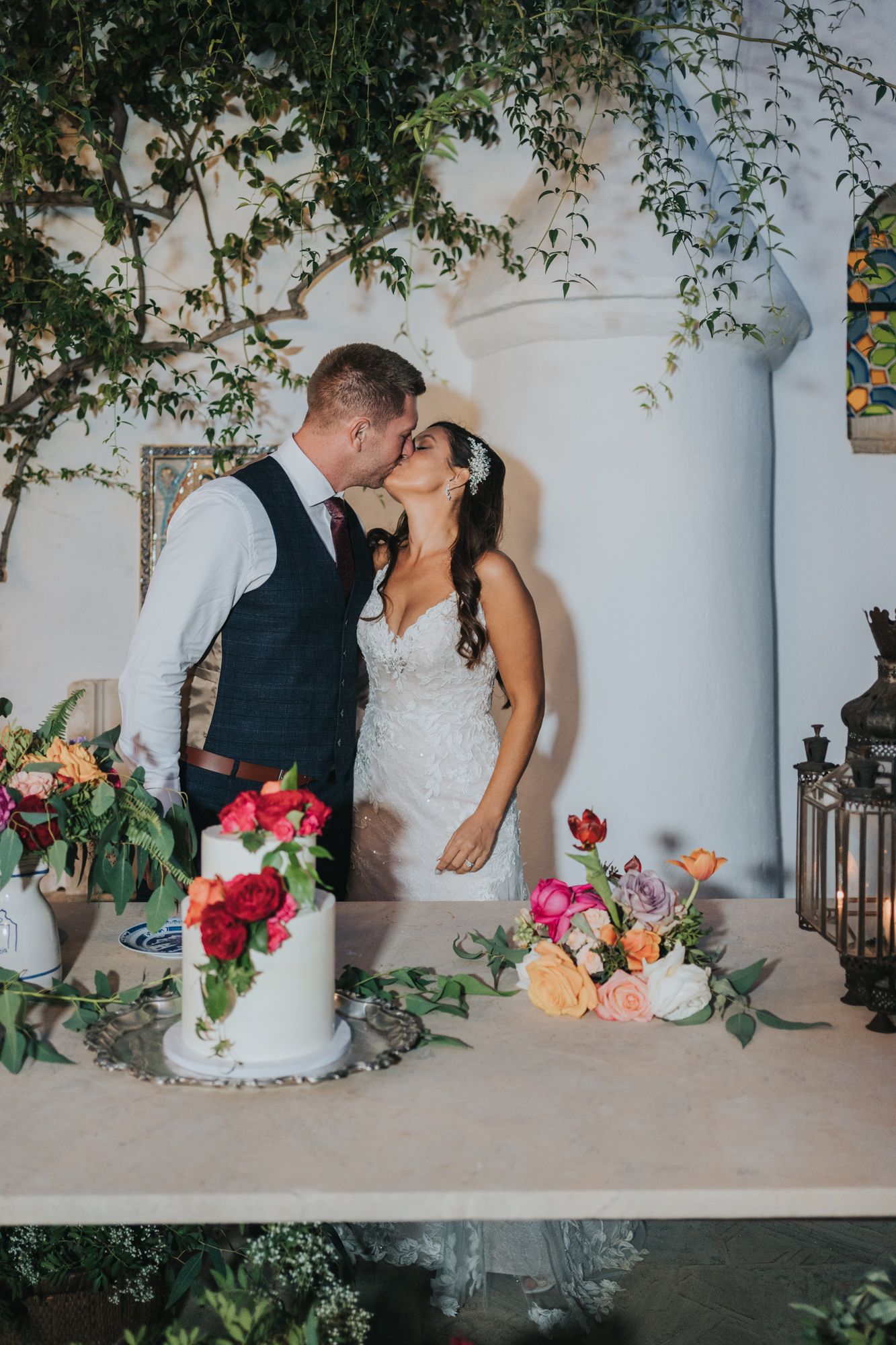 A romantic wedding scene with a couple kissing by an elegant white pillar, surrounded by floral arrangements and lanterns.