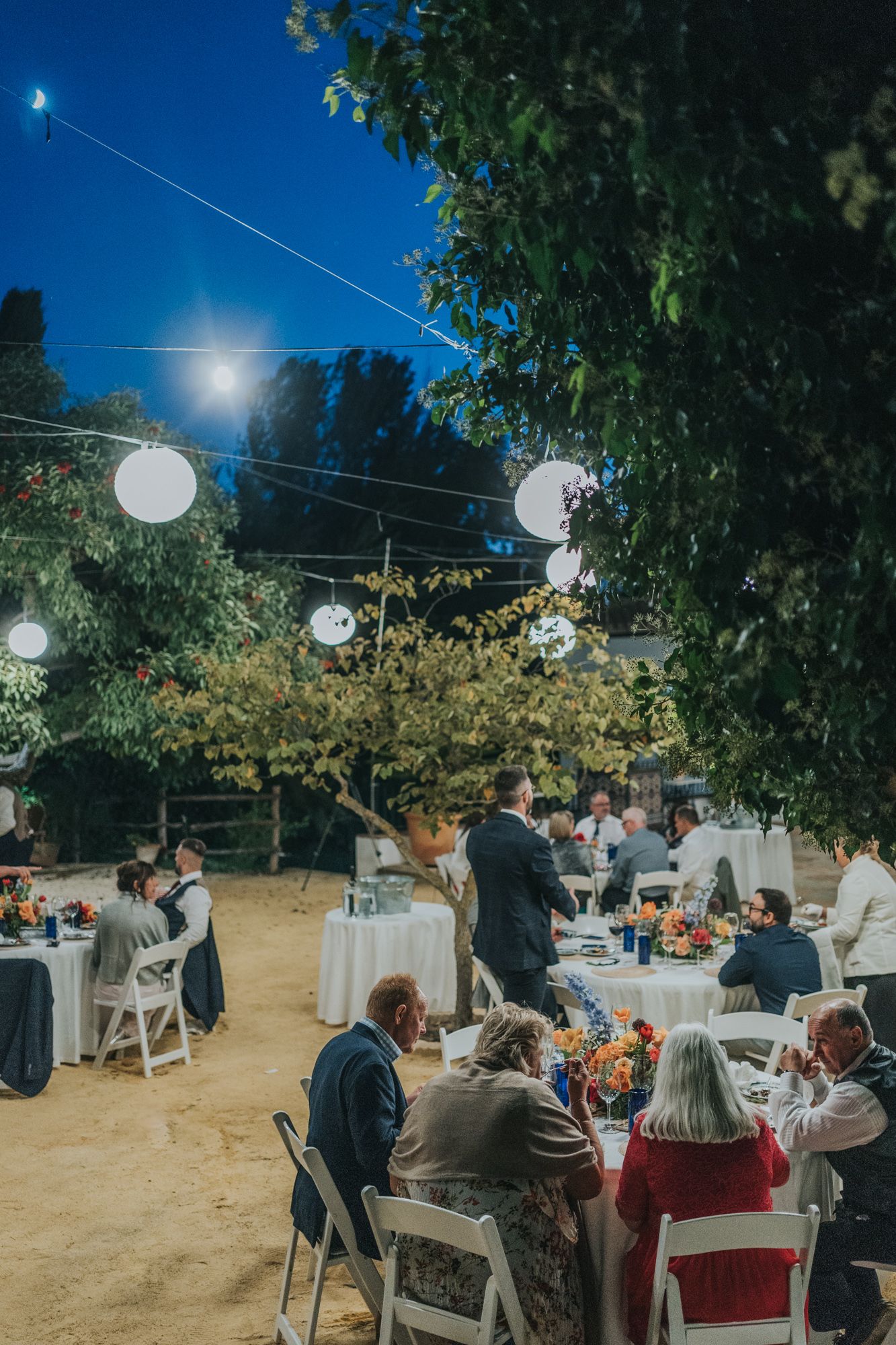Outdoor evening reception with glowing string lights and round paper lanterns illuminating tables of guests.
