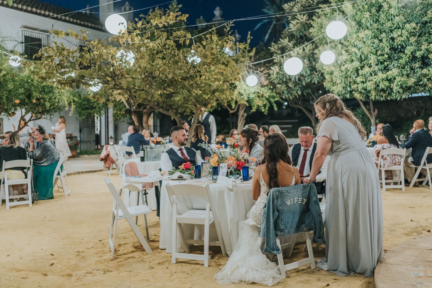 Outdoor wedding reception under string lights with guests dining at white tables in a rustic garden setting.