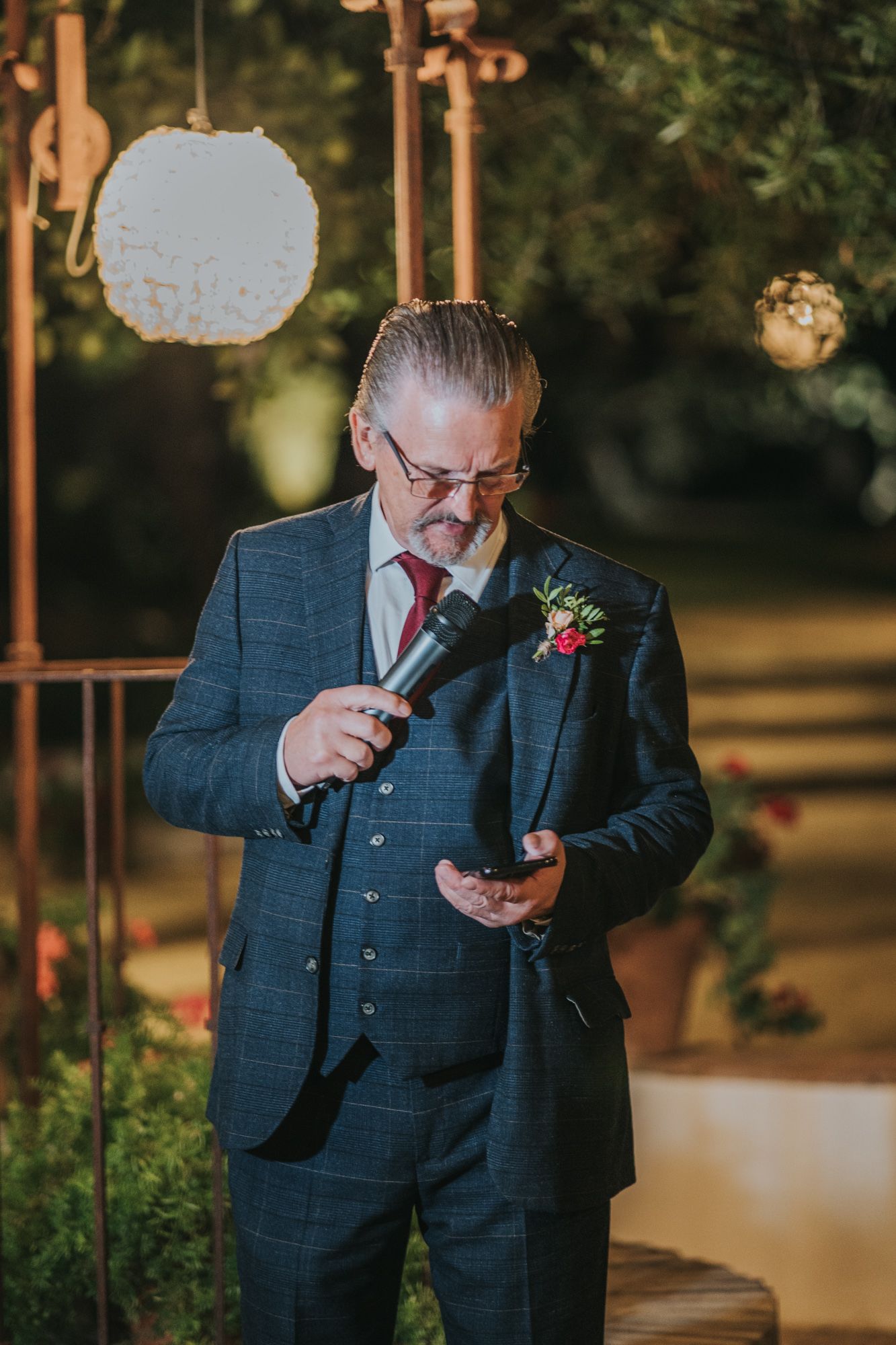 Speaker at evening outdoor event wearing navy suit and red tie holds microphone while reading notes.