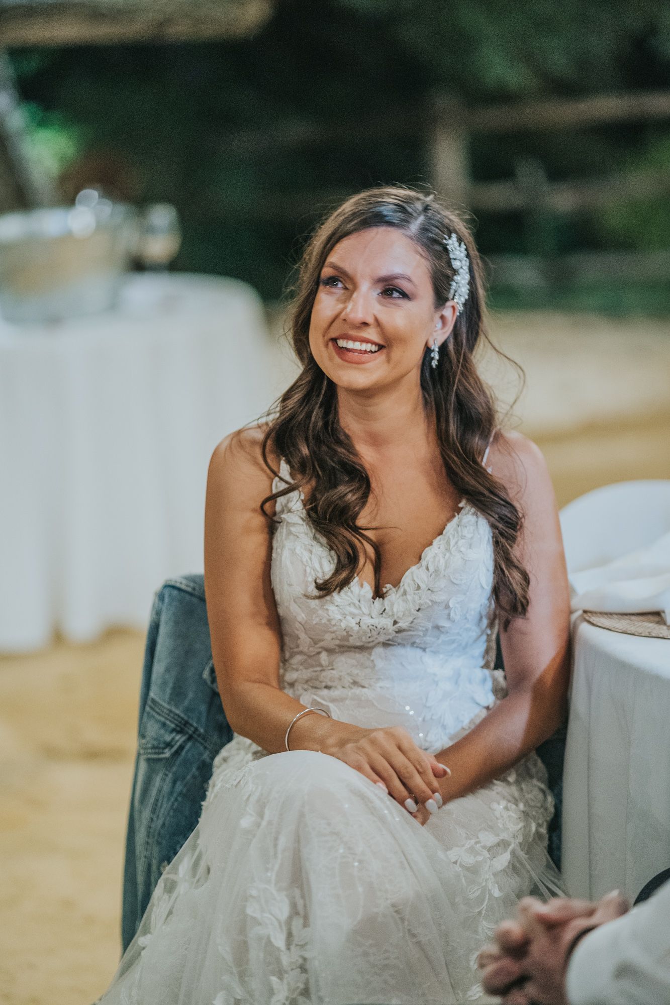 A bride in a lace wedding dress sits outdoors, smiling warmly with styled long brown hair and delicate hair accessories.