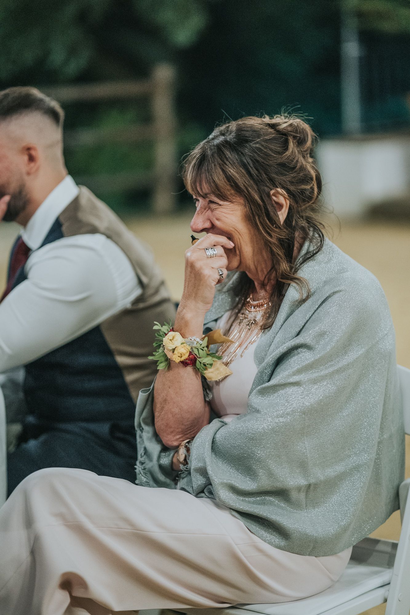 Wedding guest in grey shawl and floral corsage laughing with emotion during outdoor celebration.