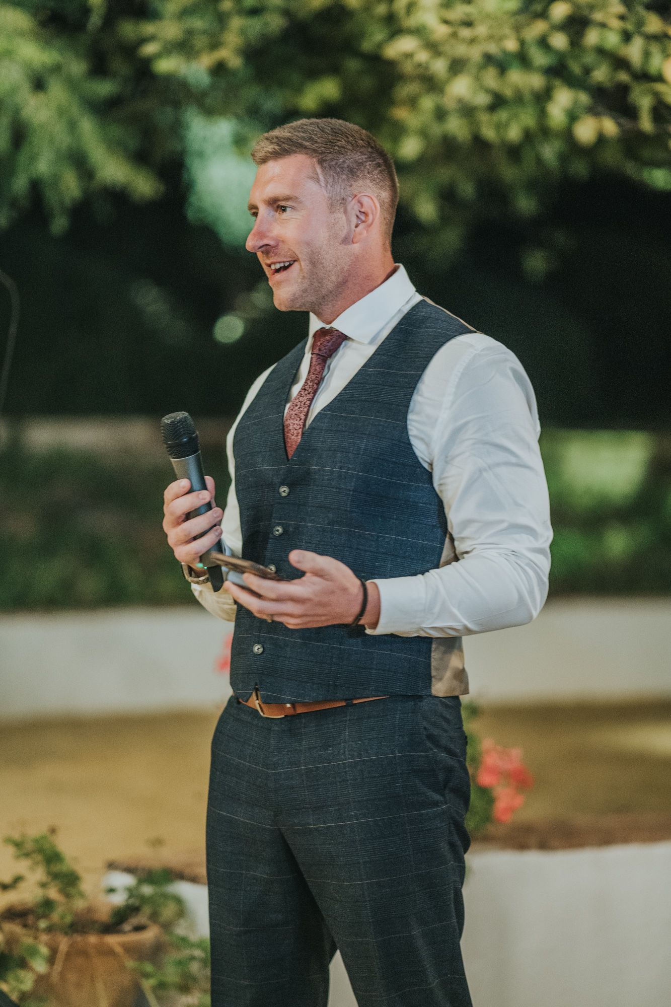 A well-dressed person in a plaid vest and tie holds a microphone while speaking at an outdoor event.