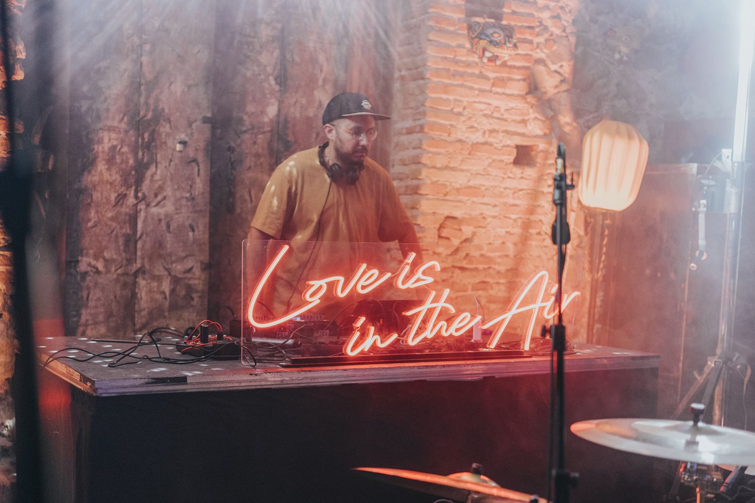 DJ mixing music behind a vintage brick wall with red neon 'Cover Band' sign illuminated in background.