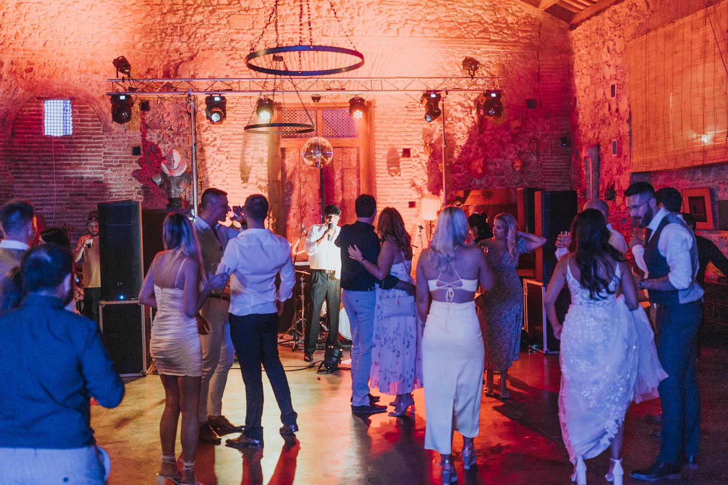 Party guests dancing in a rustic brick venue with warm red lighting and chandeliers hanging from the ceiling.