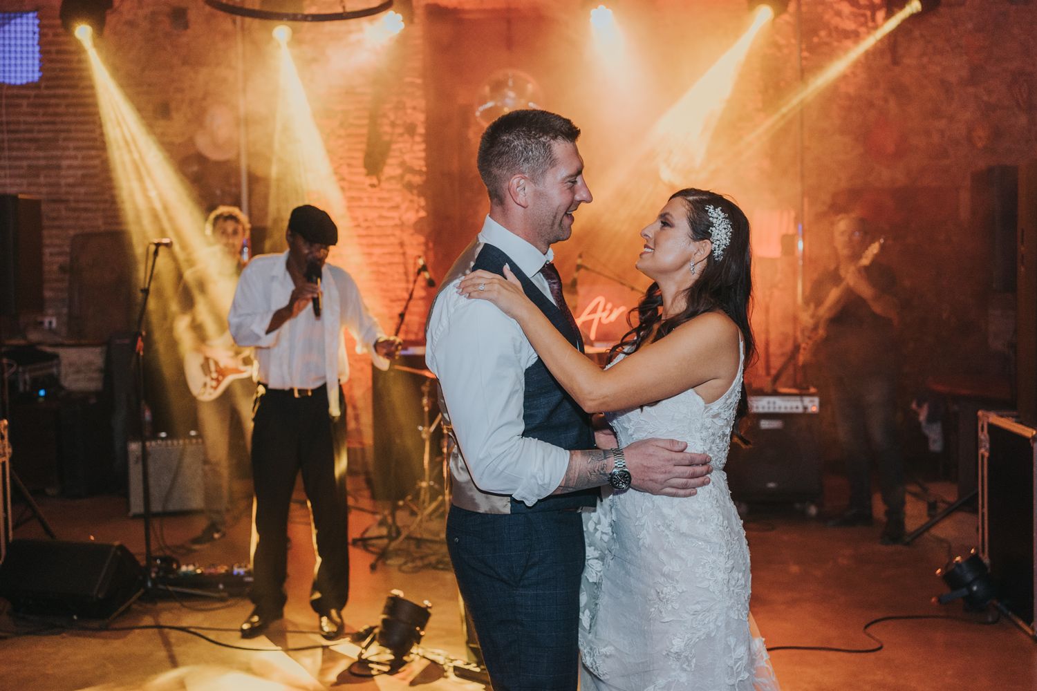 Newlyweds dancing together under vibrant orange stage lights while live band performs in background.