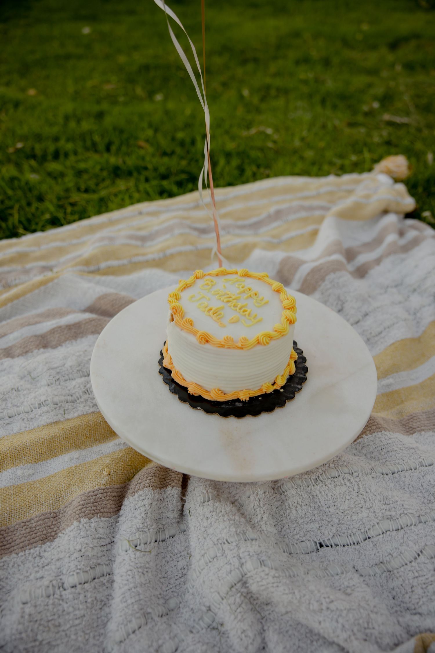 A small white birthday cake with yellow trim sits on a plate atop a striped blanket outdoors.