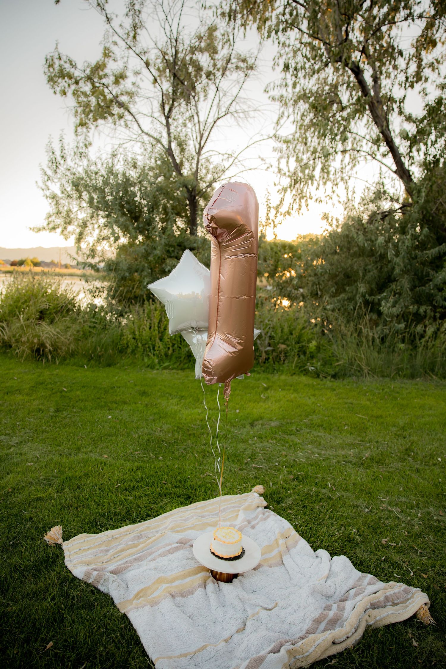 Pink balloon and silver star decorations float above a white blanket and straw hat on grass at sunset.