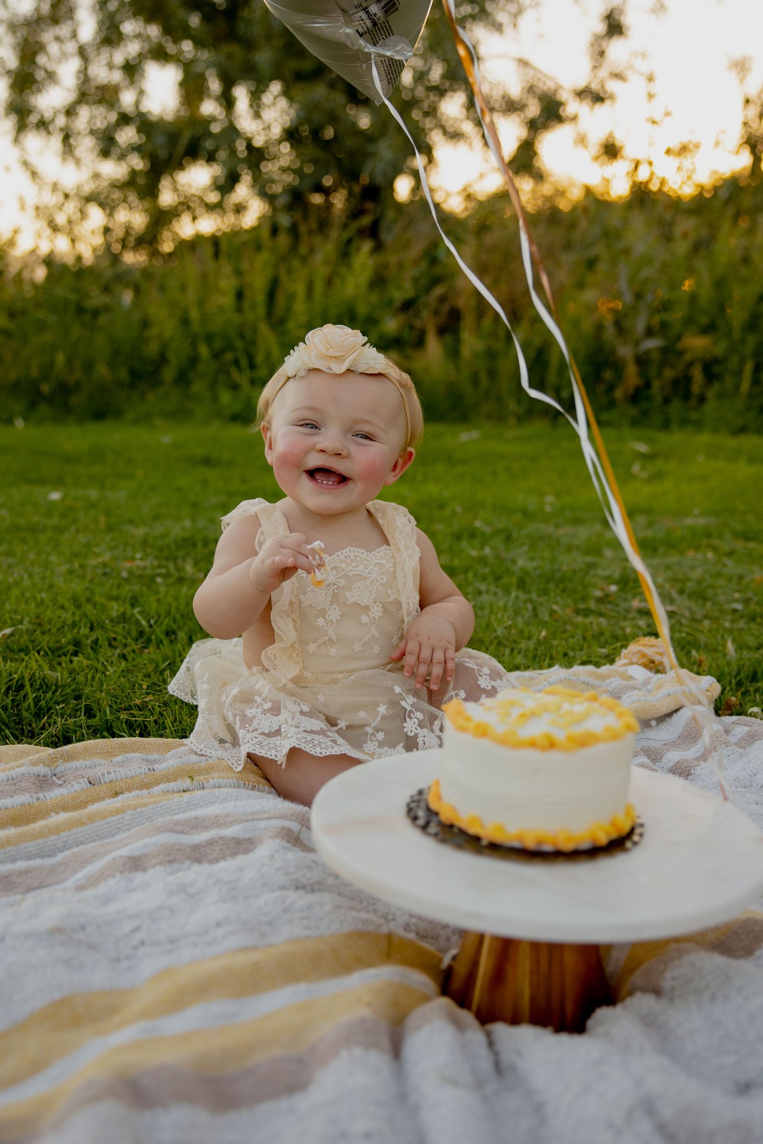 A sweet celebration setup with a yellow cake on a white blanket in a garden setting.