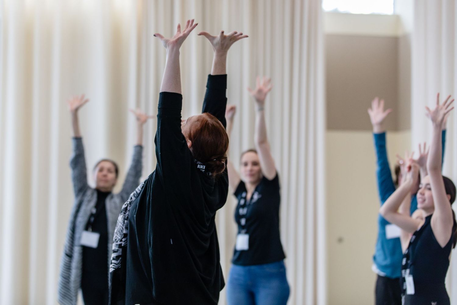 People stretching with raised arms during a group exercise or dance class in a bright studio space.
