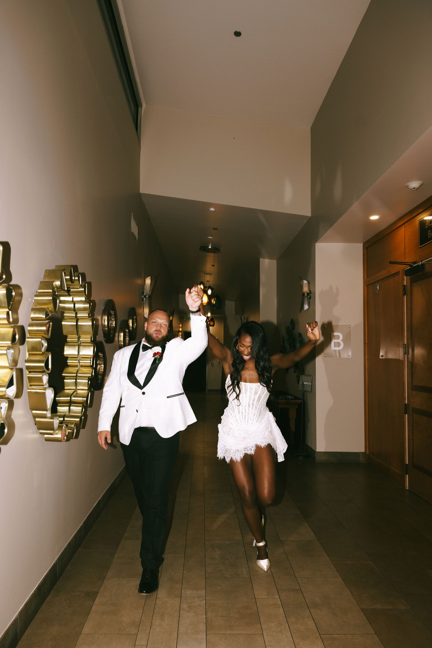 A couple in formal black and white attire dancing joyfully down a hotel hallway decorated with circular mirrors.