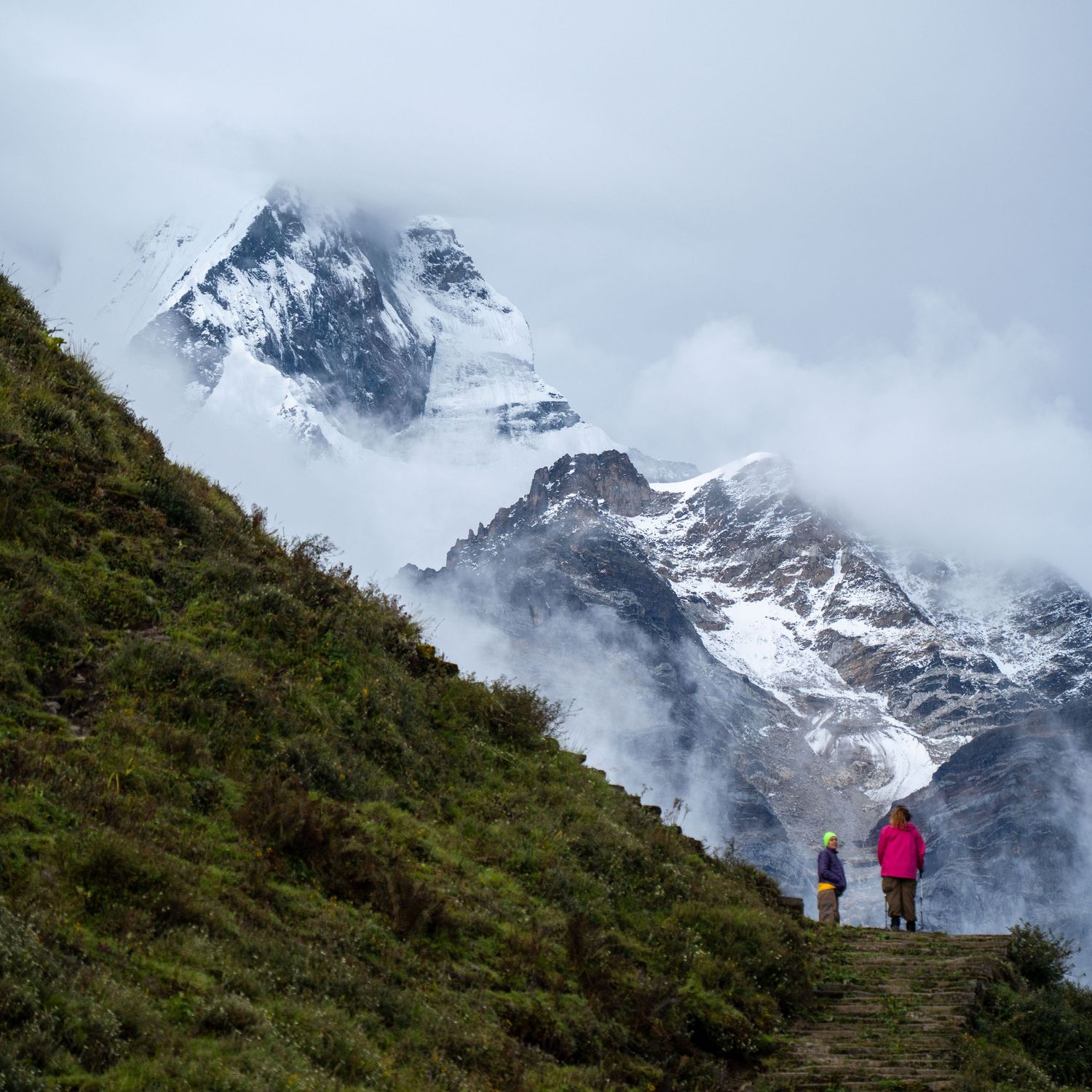 Snow-capped mountain peaks rise dramatically against cloudy skies along a winding hiking trail in Nepal.