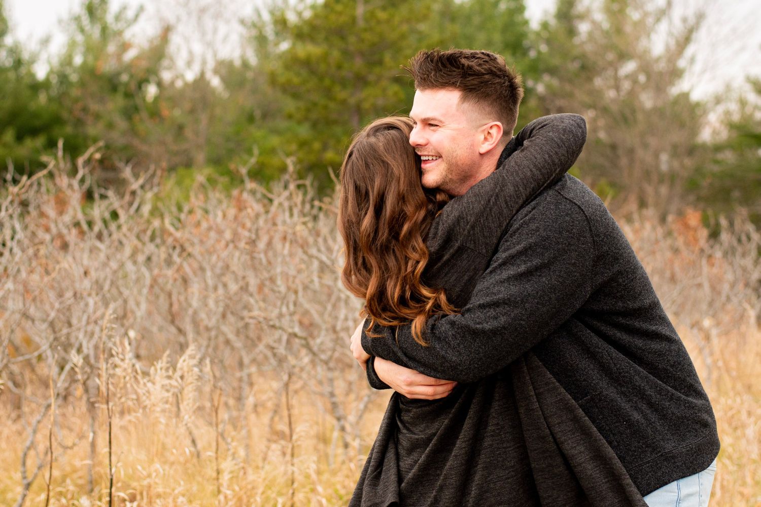 A couple embraces while sharing a romantic moment in a field of tall brown grass on an autumn day.