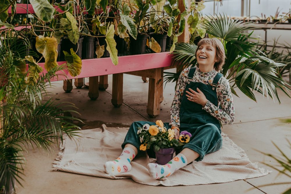 A cheerful person in a green velvet dress sits on a blanket surrounded by lush plants and yellow flowers in a greenhouse.