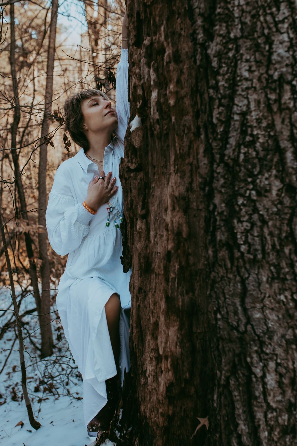 Person in white clothing leans against large tree trunk in snowy winter forest scene during golden hour.