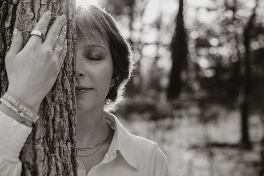 Black and white photo of someone leaning against a tree trunk with eyes closed in a peaceful forest setting.