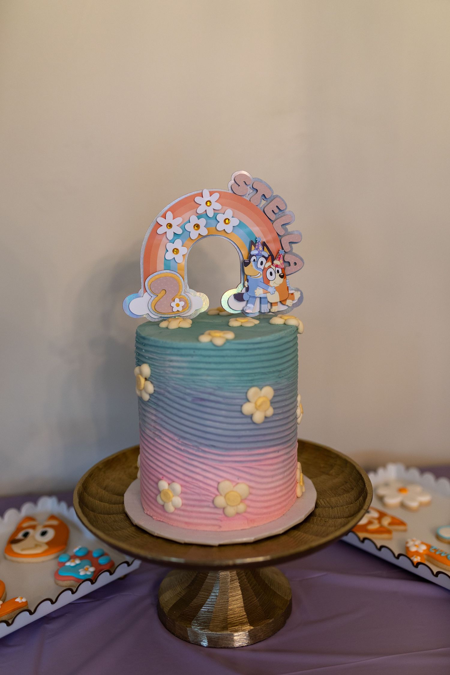 Rainbow layered birthday cake with pastel colors and Bluey topper on wooden stand.