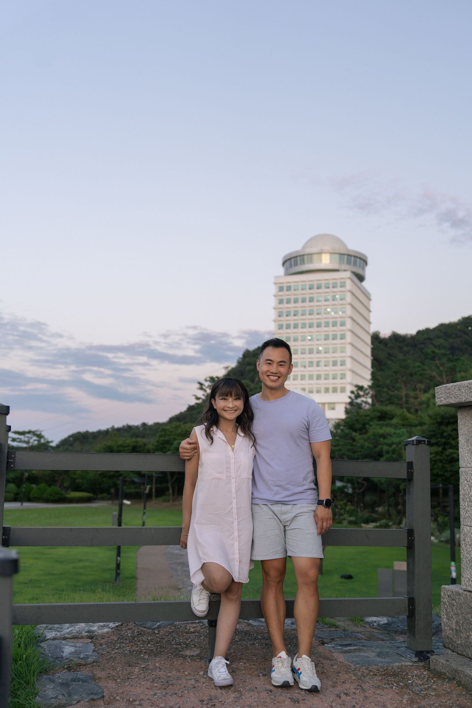 Two people stand at a scenic overlook with tall buildings and green hills in the background during sunset.