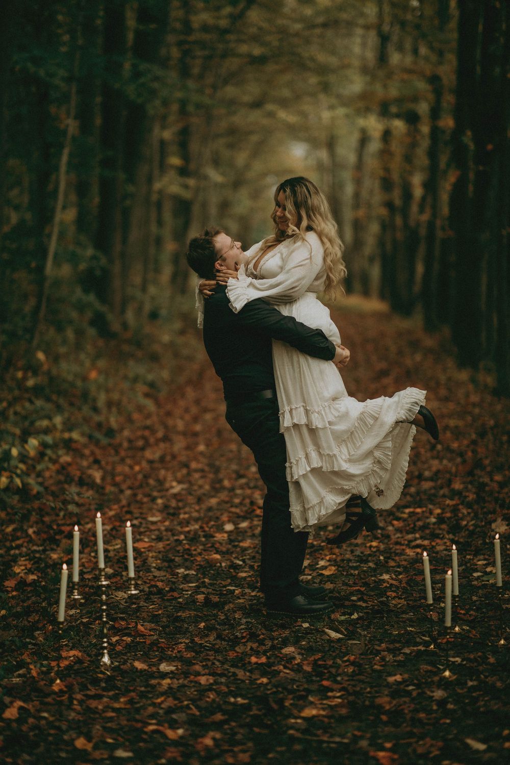 Romantic autumn forest scene with couple dancing among fallen leaves and glowing candles on the ground.
