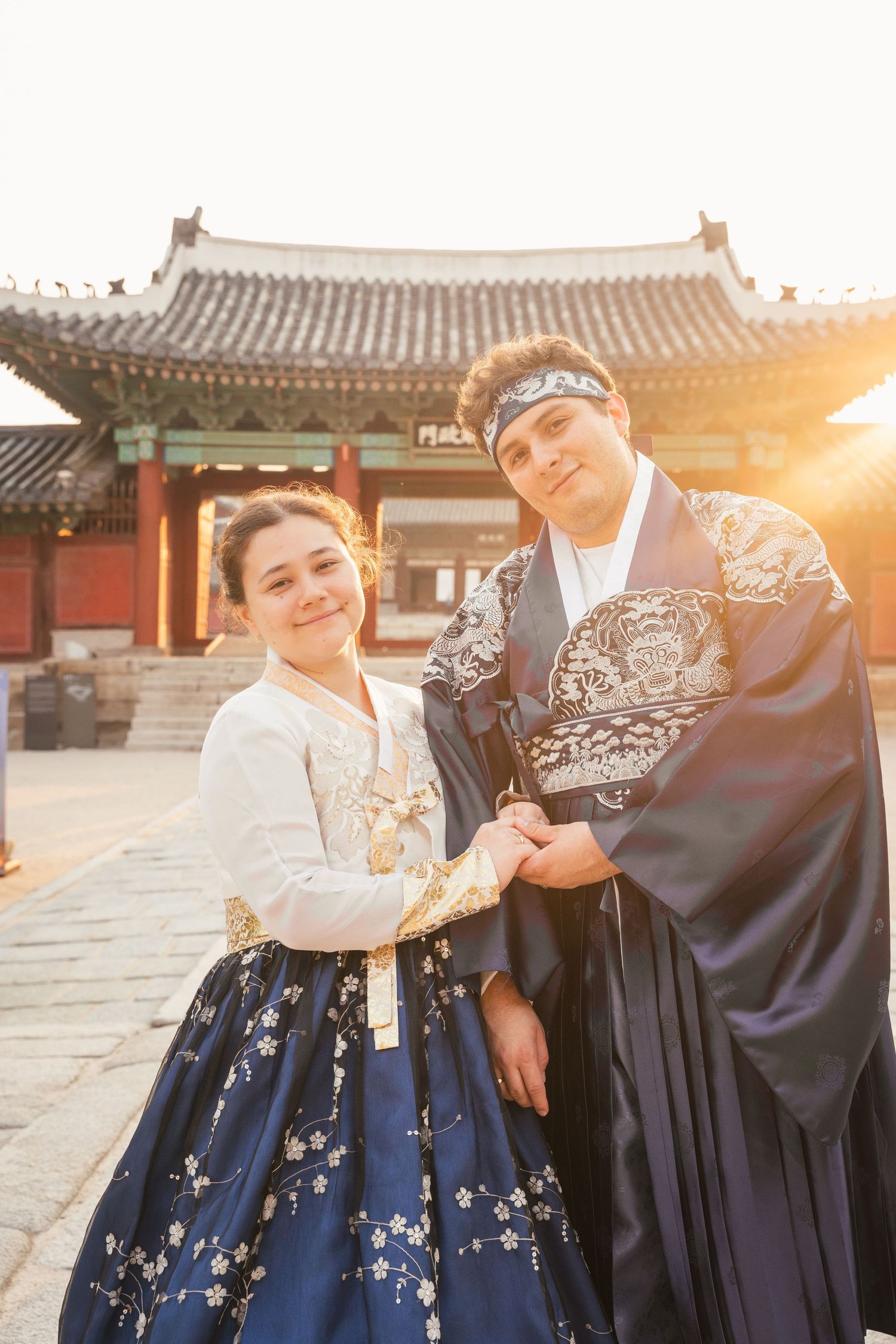 A couple in traditional Korean hanbok clothing poses together in a historic palace courtyard during sunset.