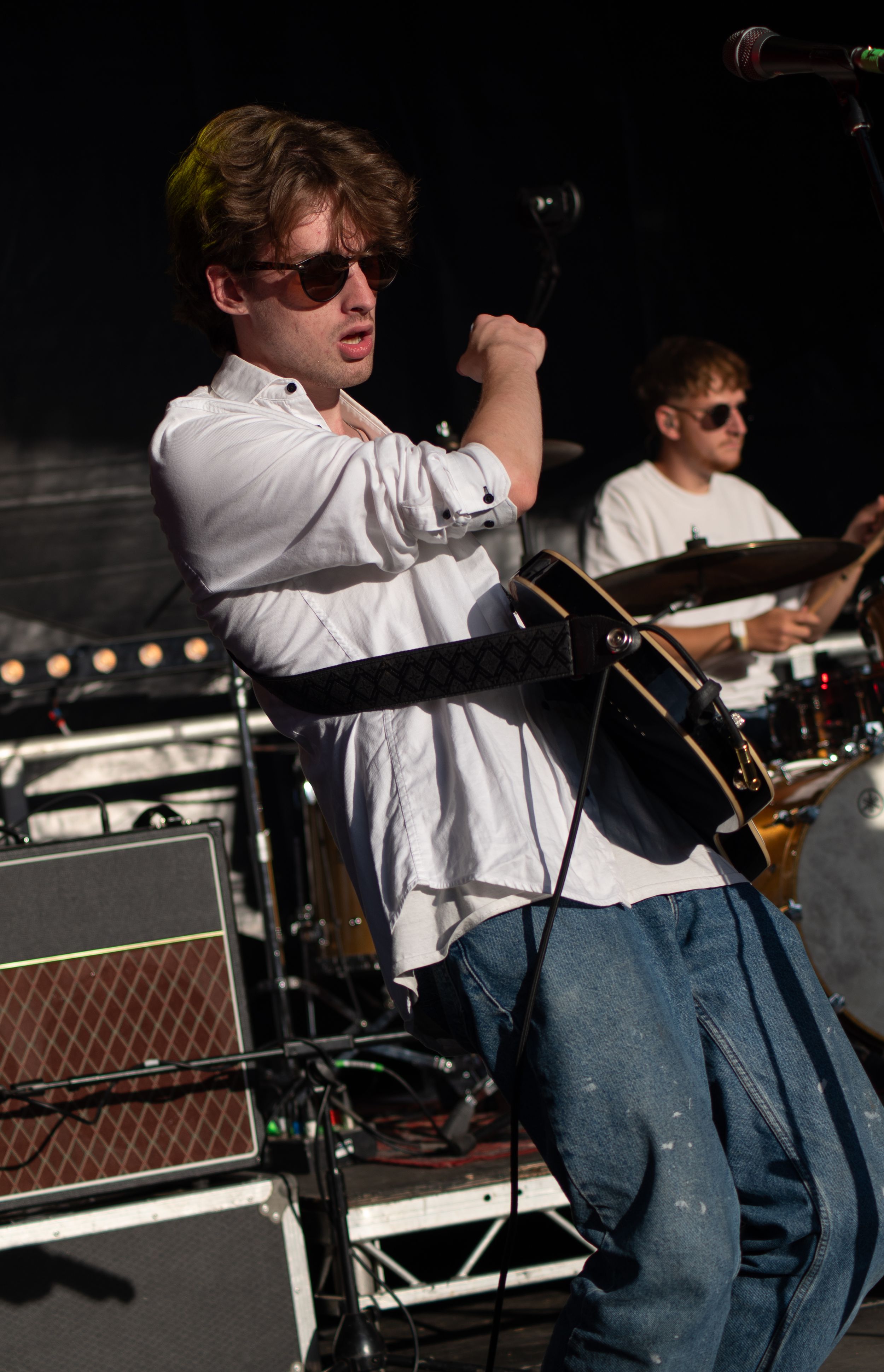 Musician in white shirt and jeans performs energetically with guitar on outdoor stage during daytime concert.