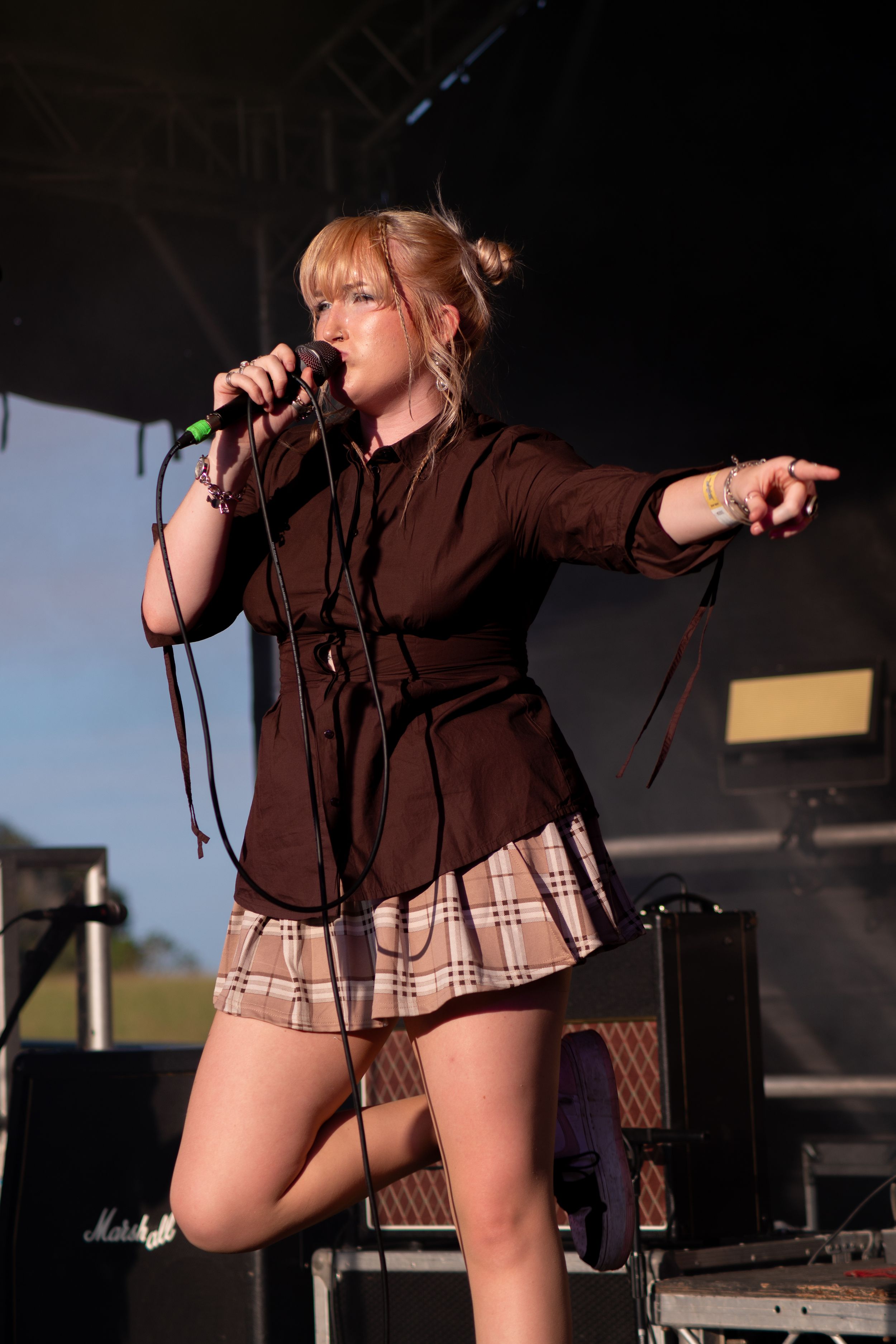 A performer in a brown top and plaid skirt sings energetically on stage during a live concert performance.