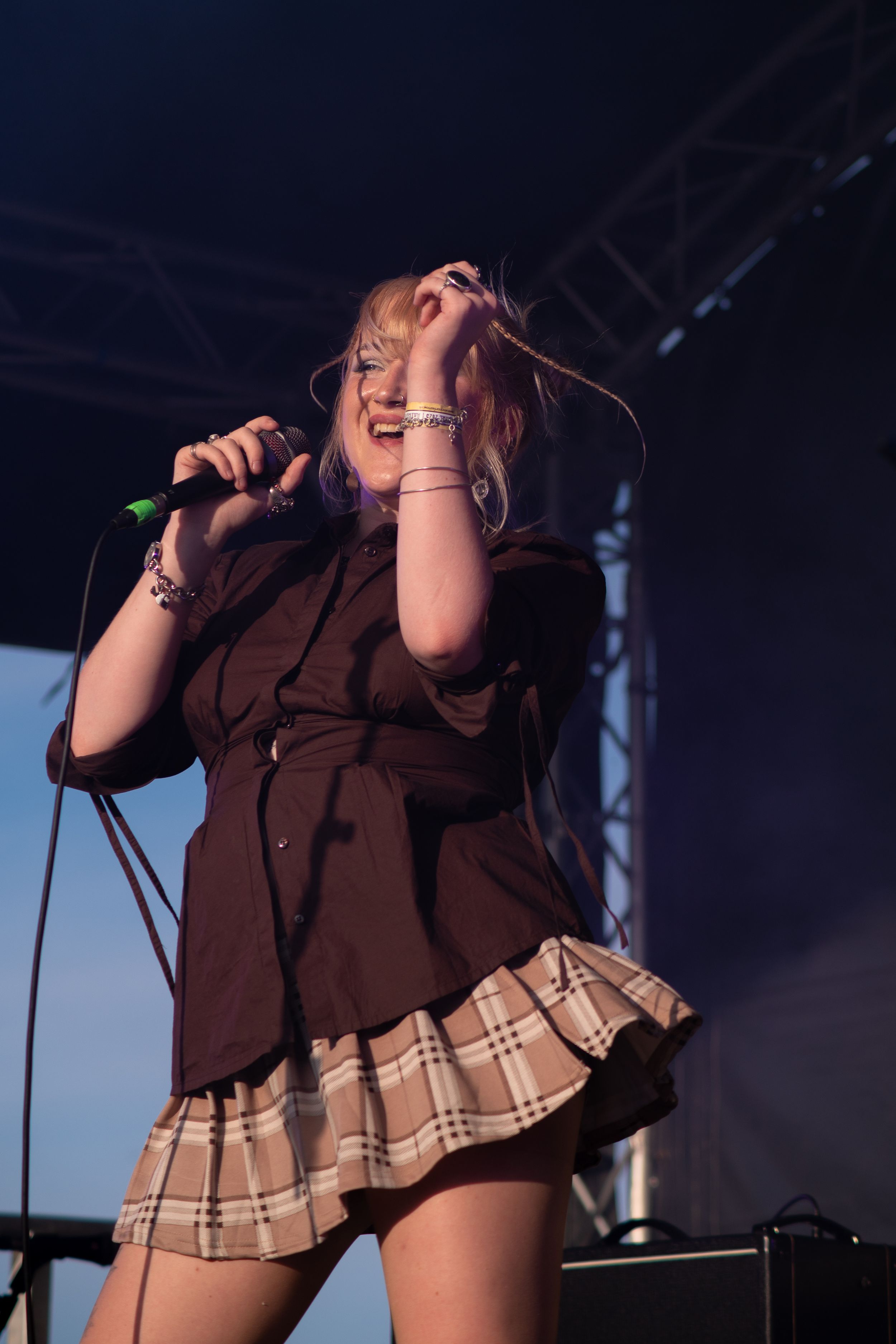 A performer in a brown top and plaid skirt sings passionately on stage during a live music performance.