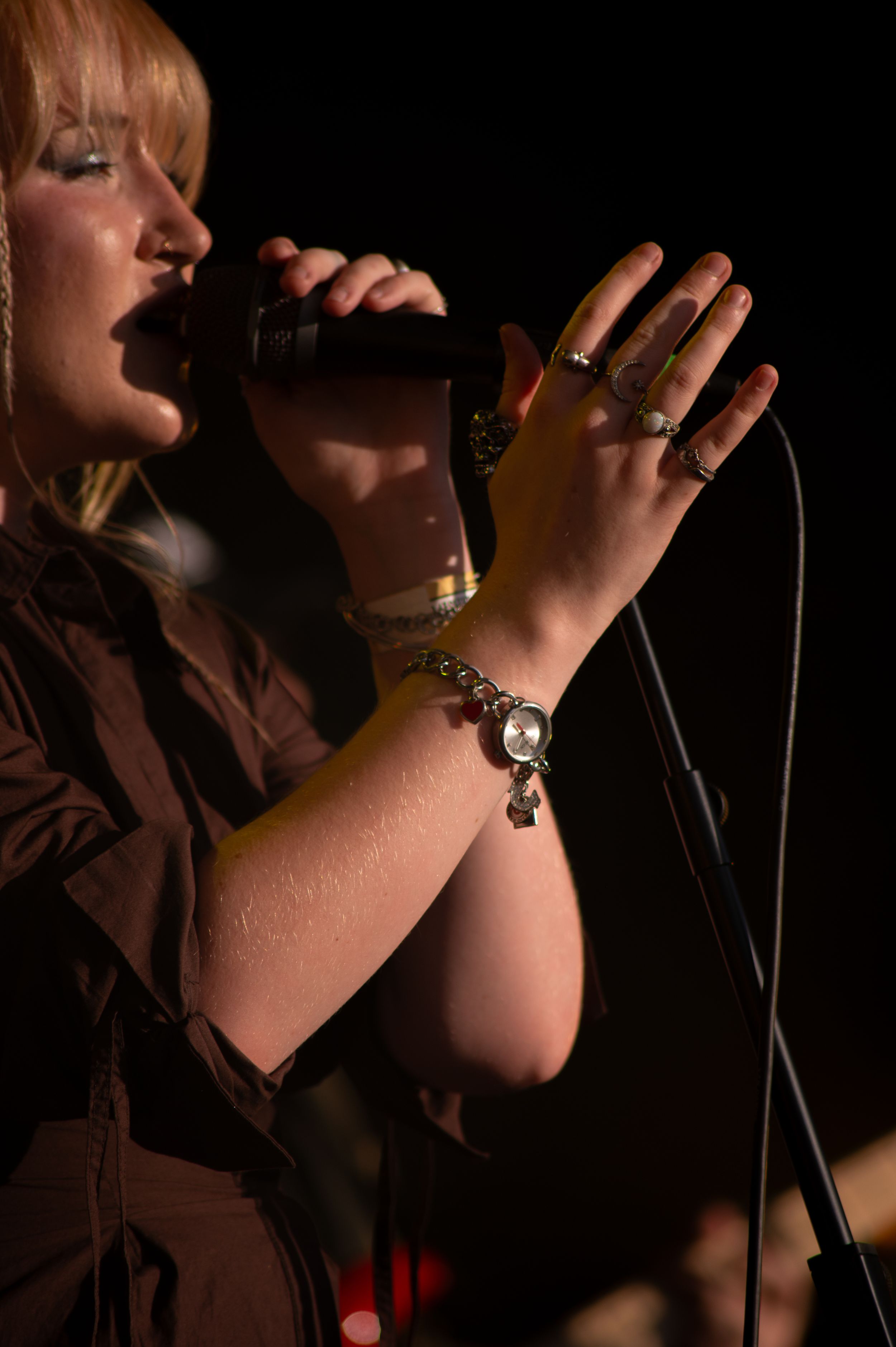 A singer performs on stage in dramatic lighting while wearing a bracelet and rings, holding a microphone close to their mouth.