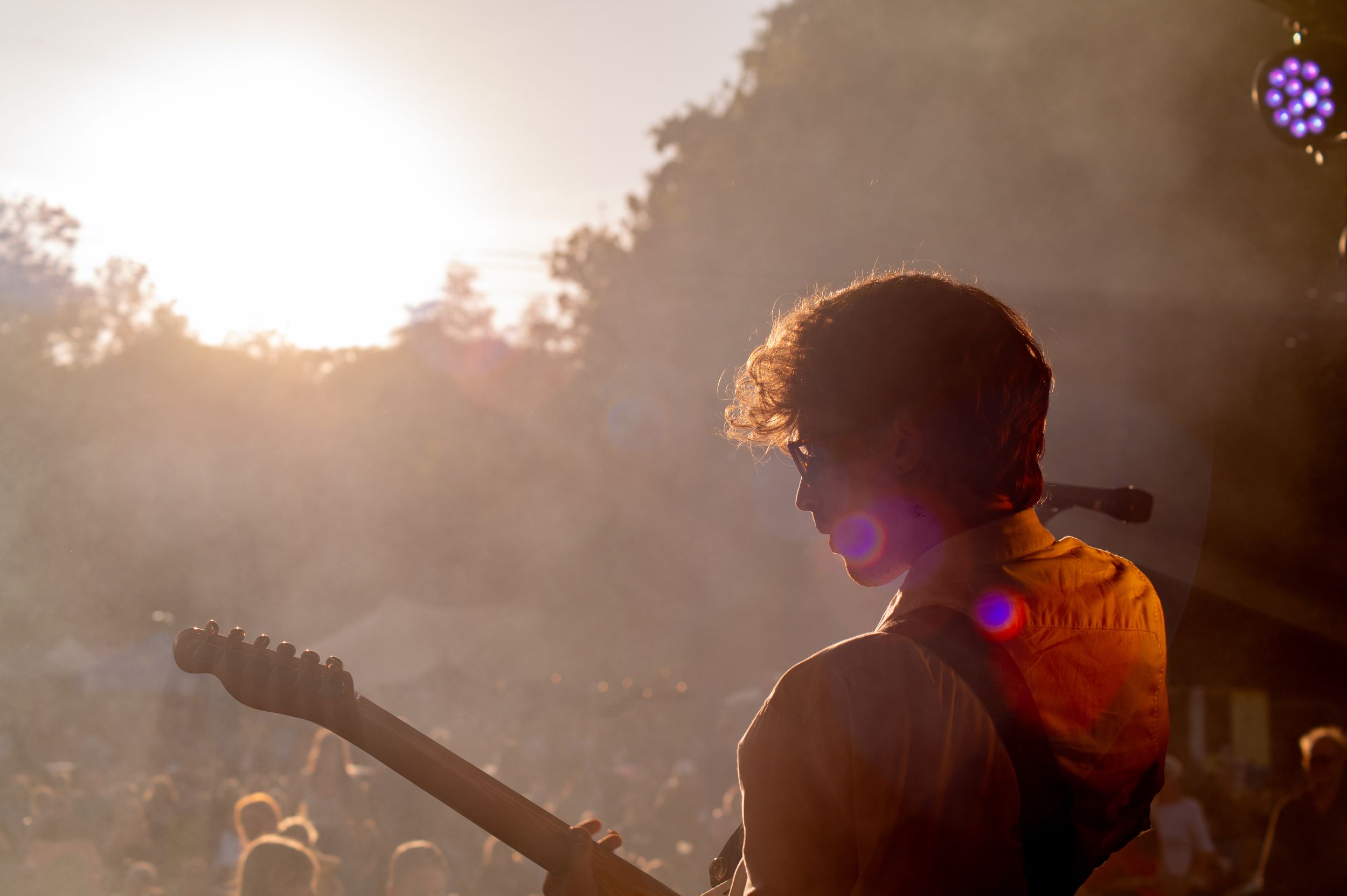 Silhouetted guitar player performs on stage during sunset with warm golden lighting and lens flare effects.
