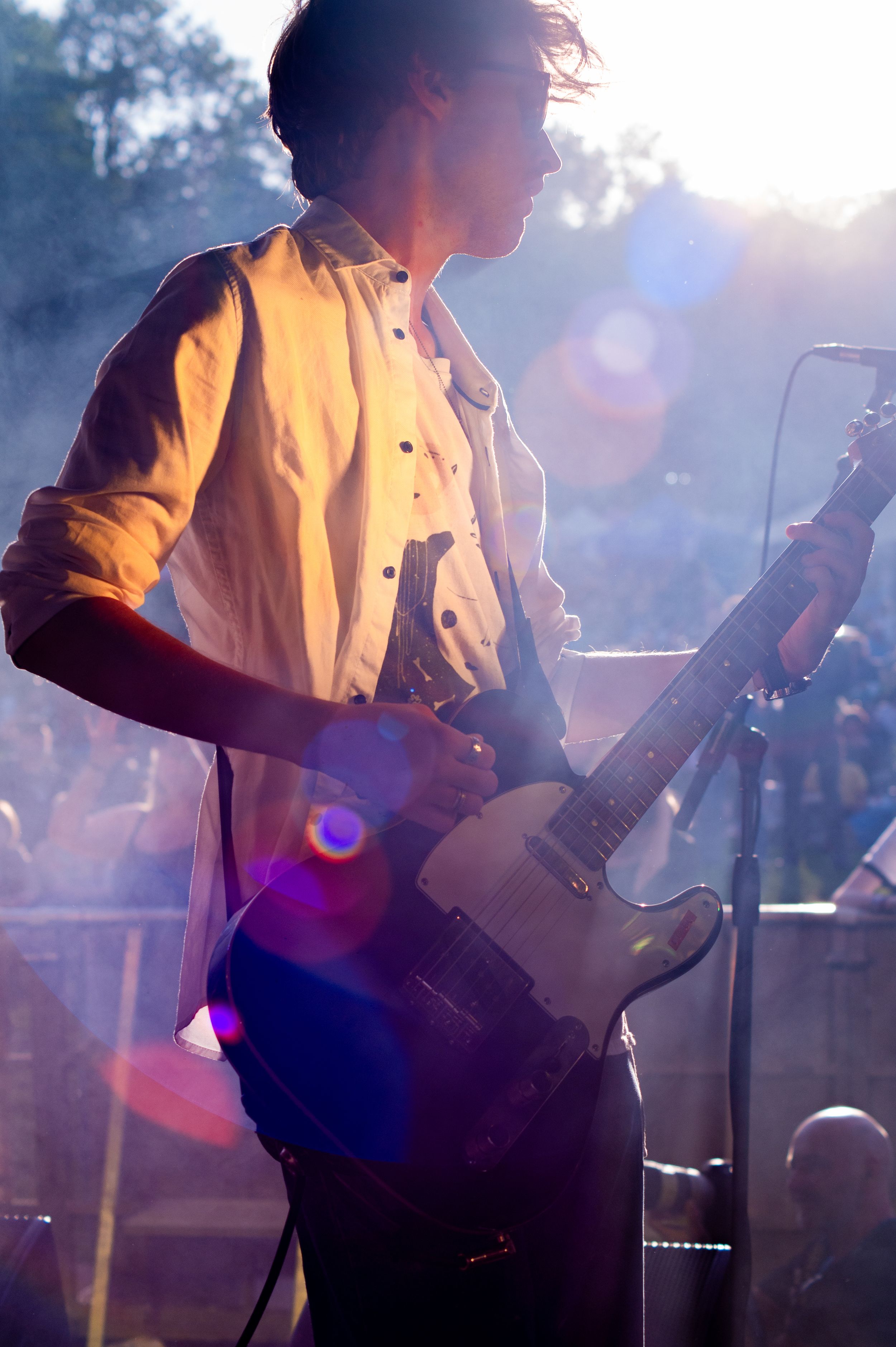Guitarist performs on stage in white shirt with dramatic backlighting and lens flare effects during live concert.