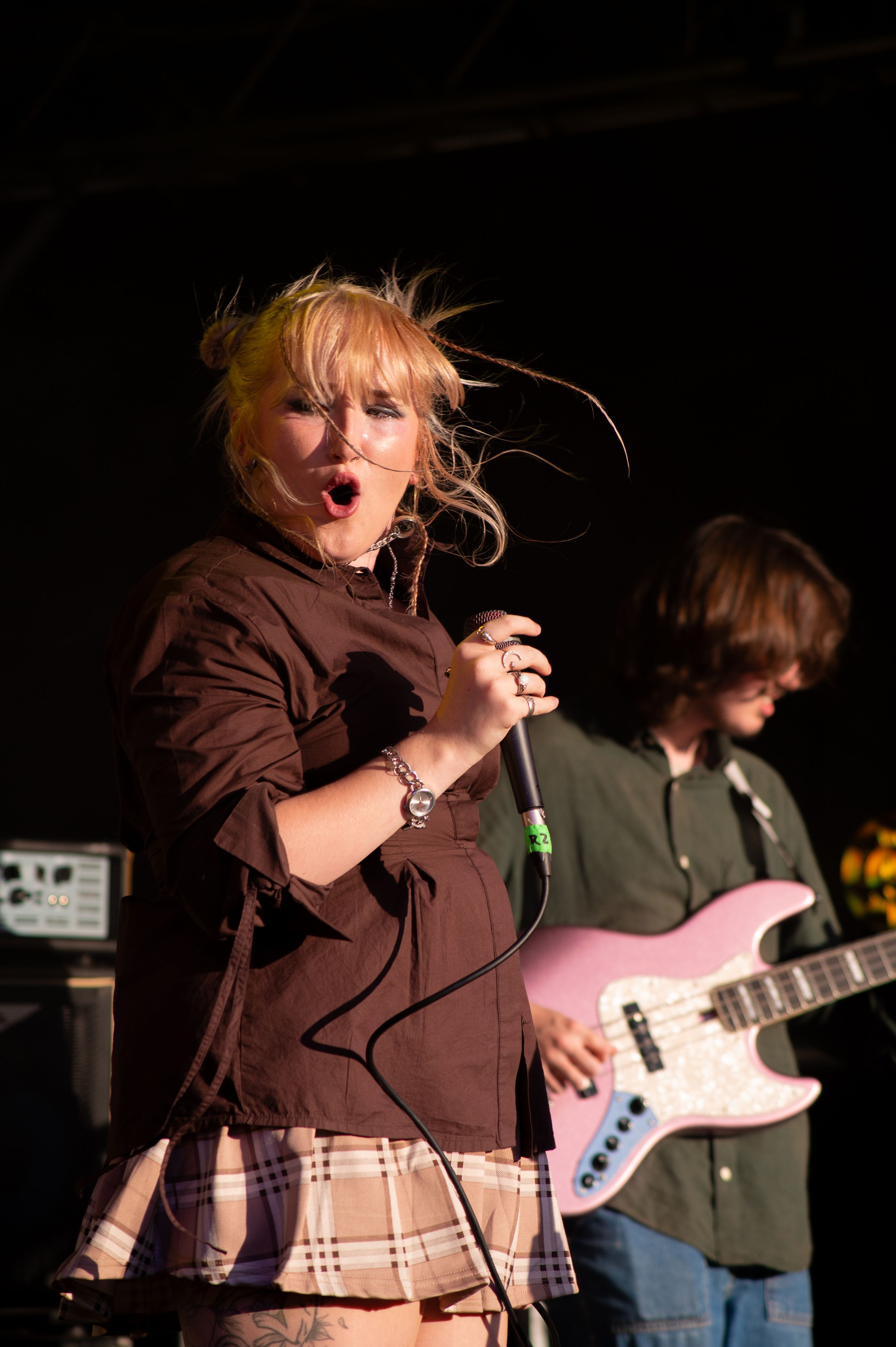 Performer sings passionately into microphone wearing brown top and plaid skirt while bassist plays in background.