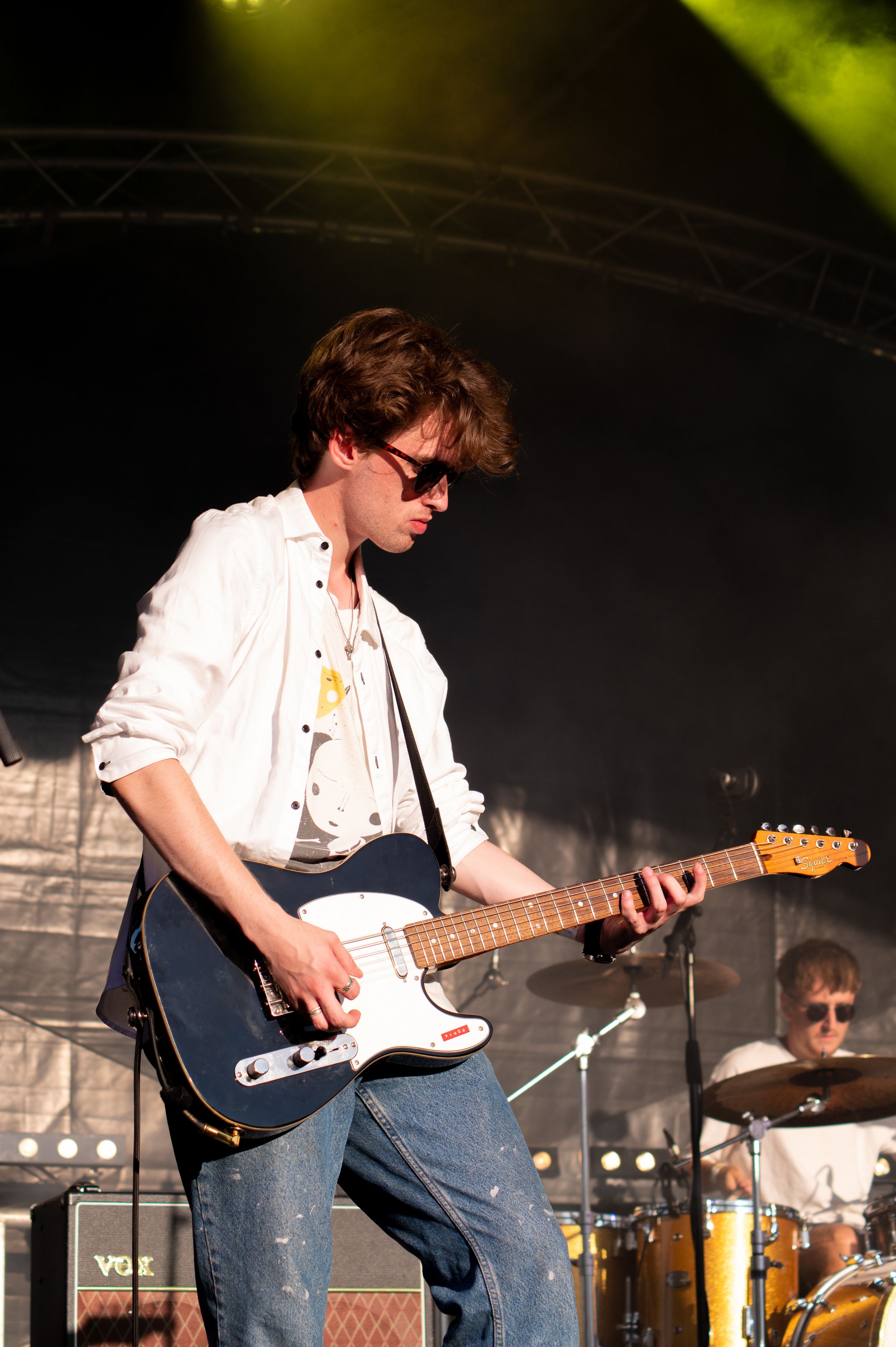 Guitarist in white jacket and sunglasses plays Fender Stratocaster on outdoor festival stage with yellow lighting.