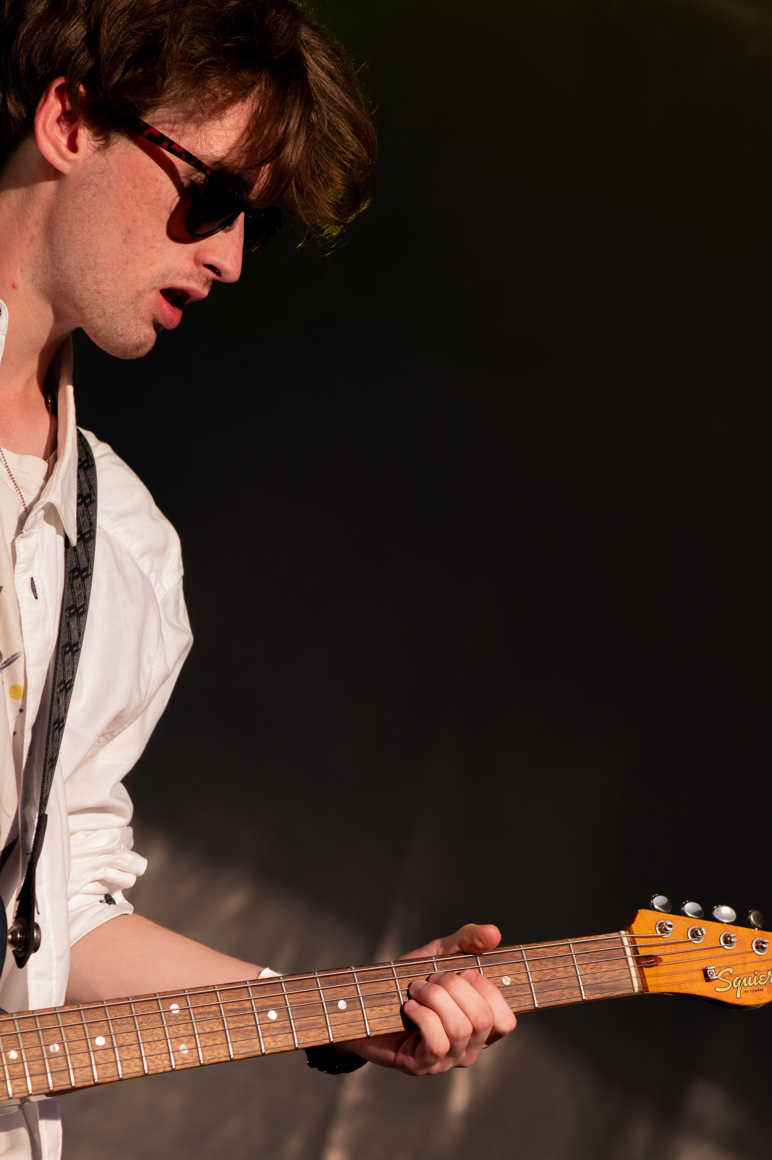 A musician in sunglasses and white shirt plays an electric guitar on stage against a dark background.