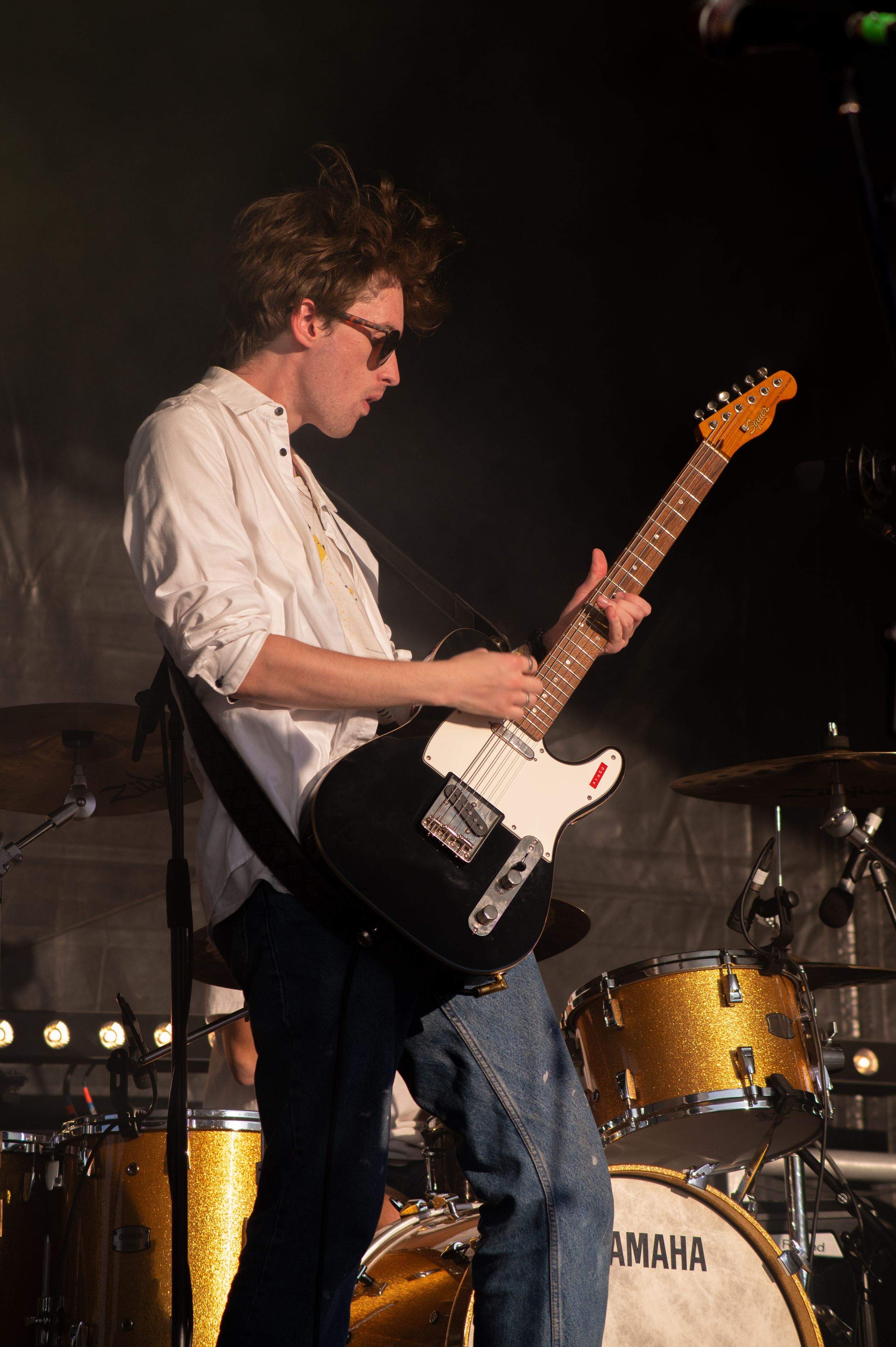 Guitarist in a white shirt performs on stage with a Yamaha drum kit visible in the background.