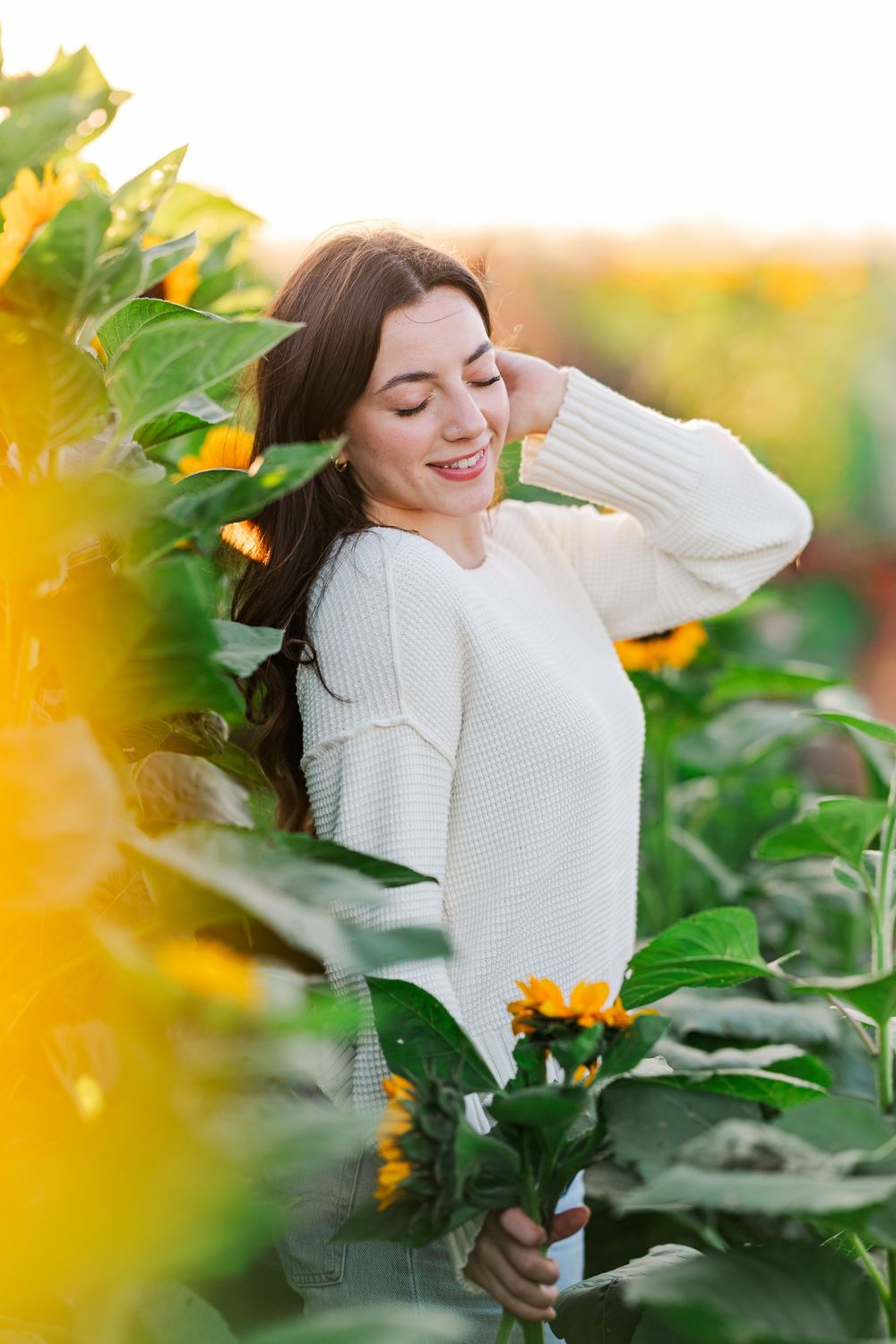 Woman in white sweater enjoys autumn sunlight among yellow flowers and green foliage in garden setting.