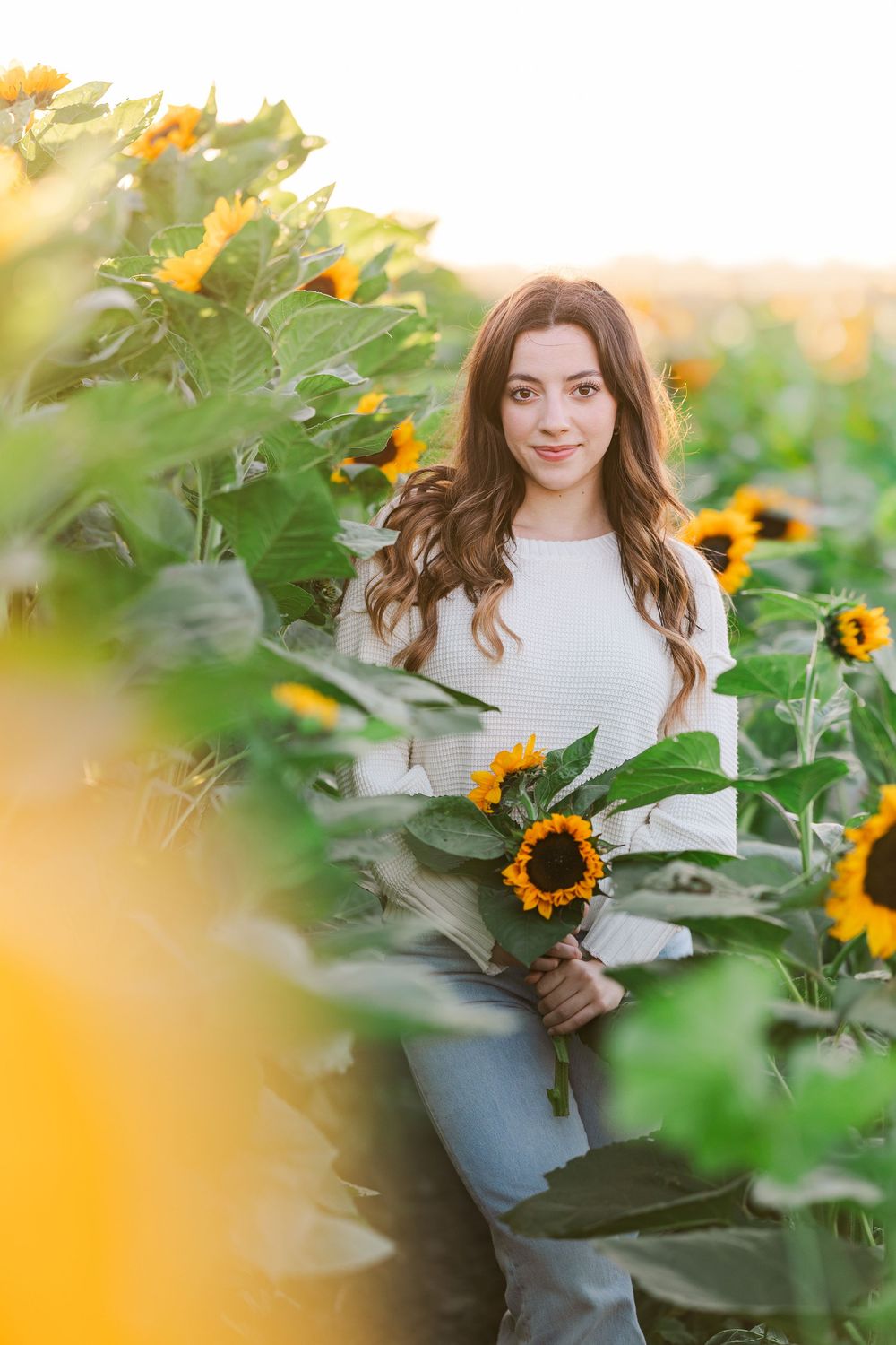 Person in white sweater poses joyfully among vibrant sunflowers in a garden during golden hour.