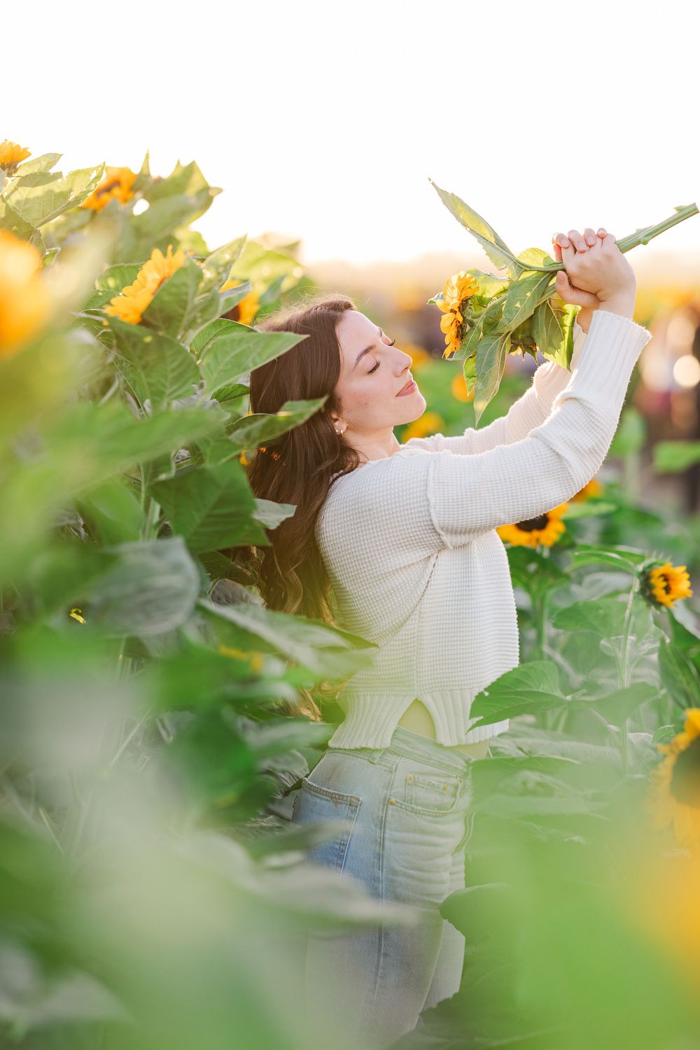 Person in white sweater dances carefree among tall sunflowers at sunset with arms raised in celebration.