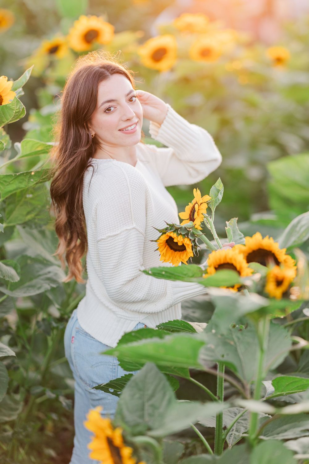 A person in a white sweater poses joyfully among bright yellow sunflowers in a garden during golden hour.