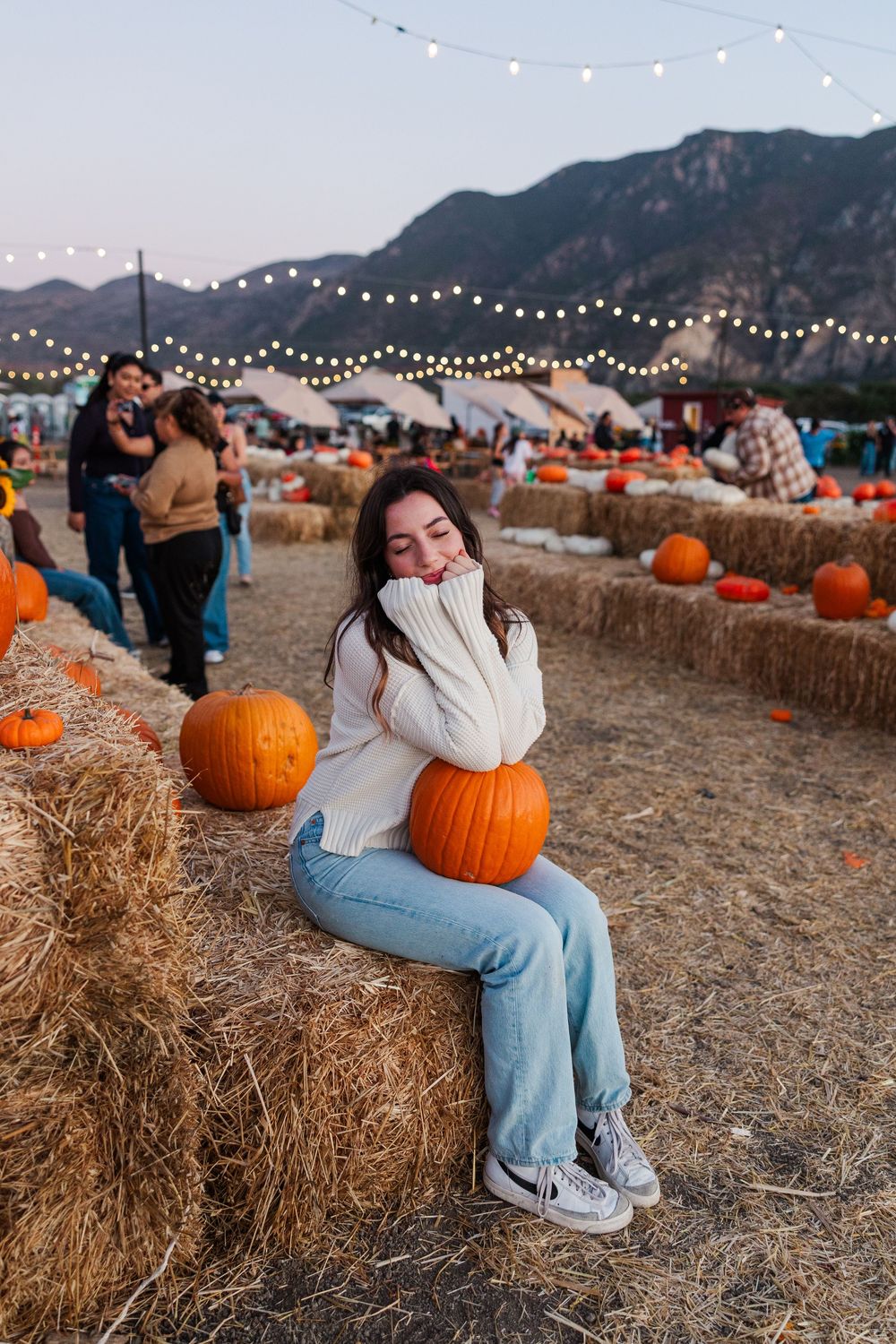 A pumpkin patch at sunset with hay bales, string lights, and mountains in the background creates a cozy autumn scene.