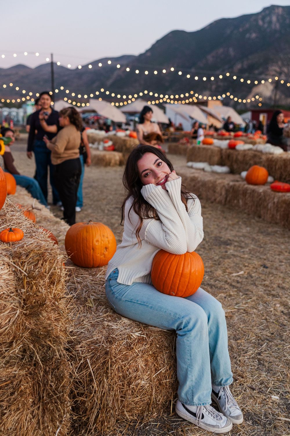 String lights illuminate a rustic pumpkin patch with hay bales and mountains at dusk.