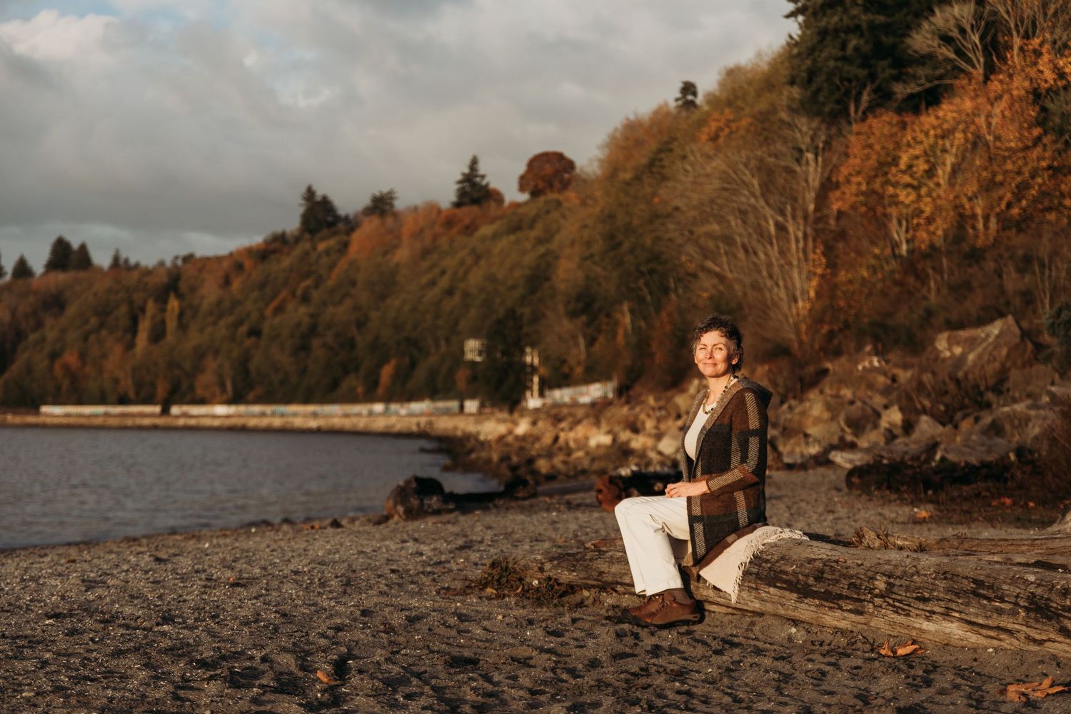A couple embraces on a rocky beach at sunset with autumn foliage and mountains in the background.