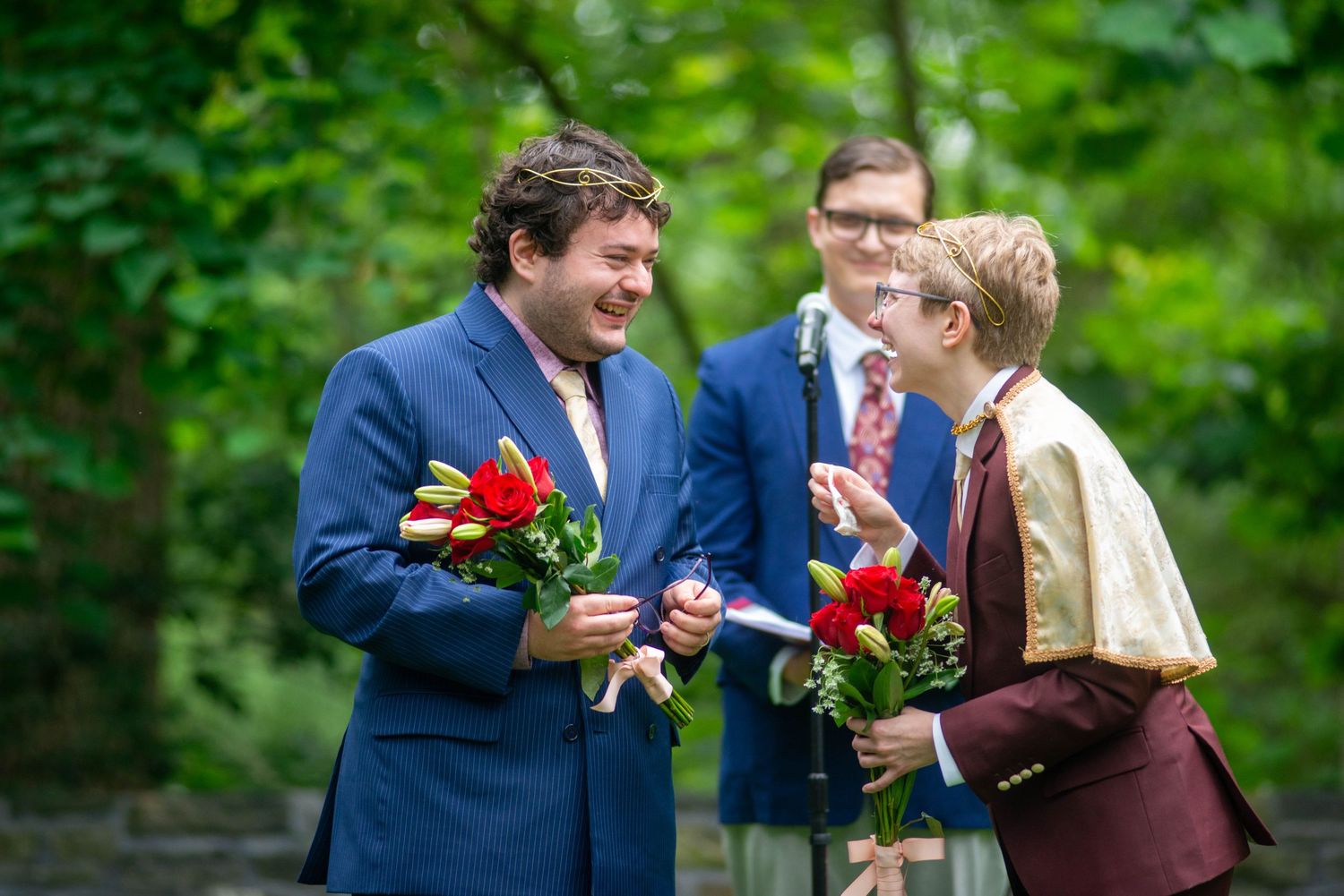 A couple shares a tender moment in a garden setting while holding red roses and wearing formal attire.