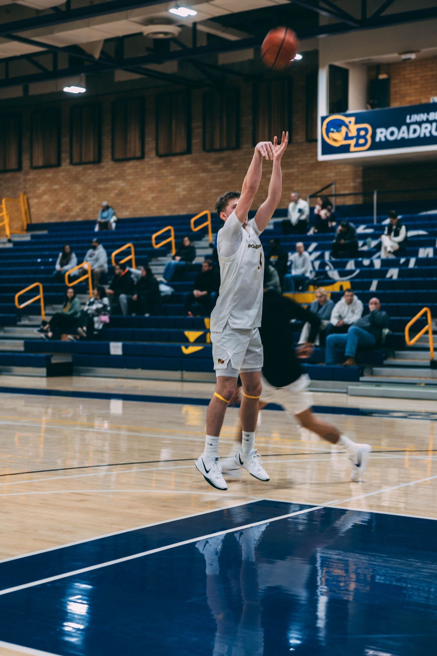 Basketball player takes a jump shot on an indoor court with bleachers in background.