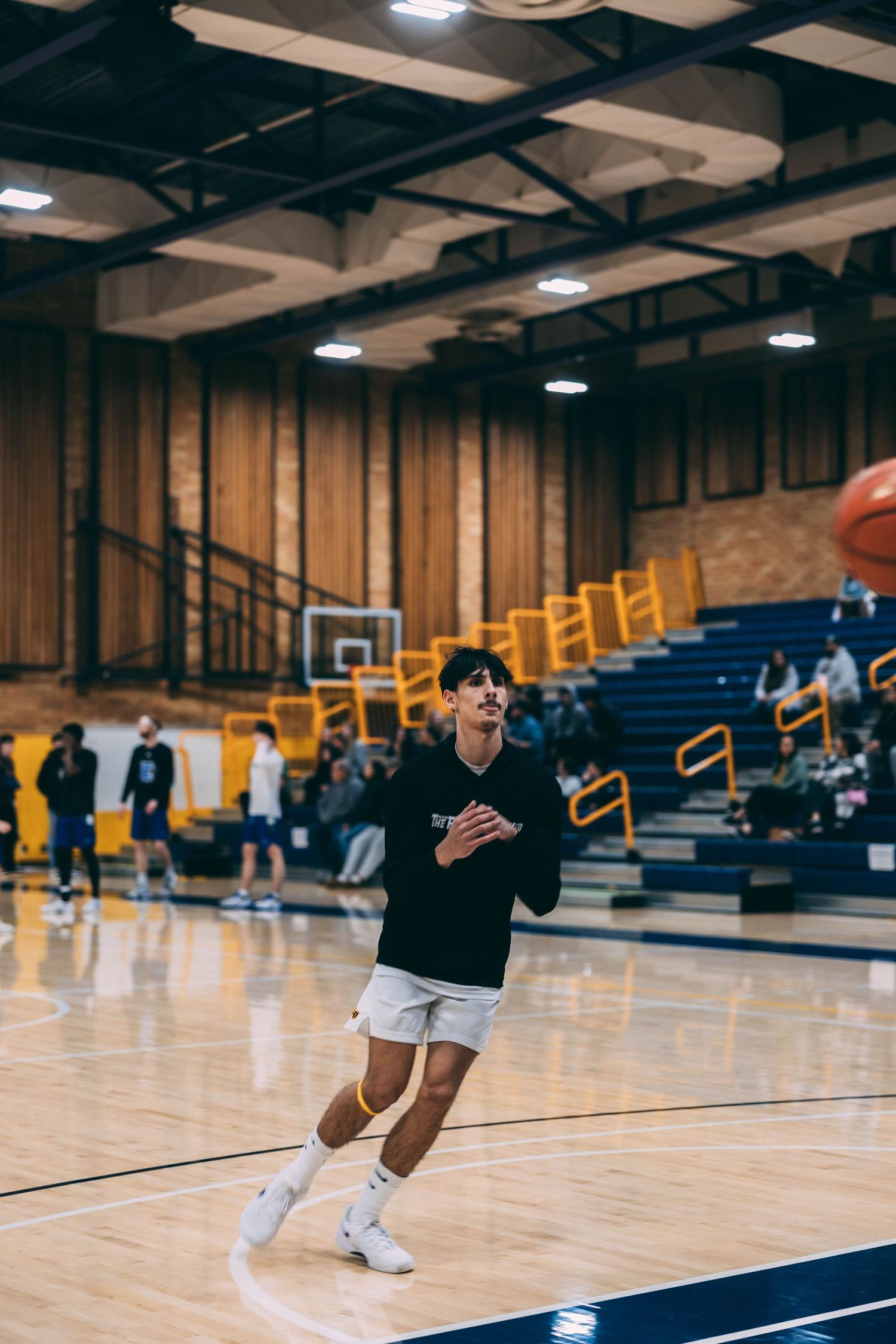 Basketball player in black shirt and white shorts practices on indoor court with blue bleachers in background.