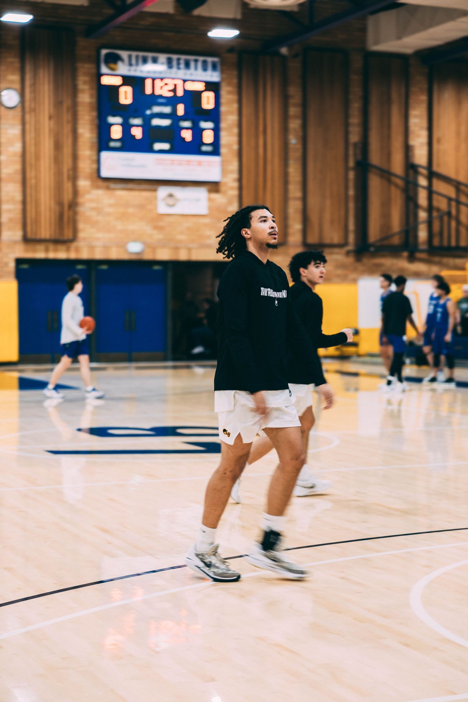 Player jumps during basketball practice on polished gymnasium floor with blue bleachers visible.