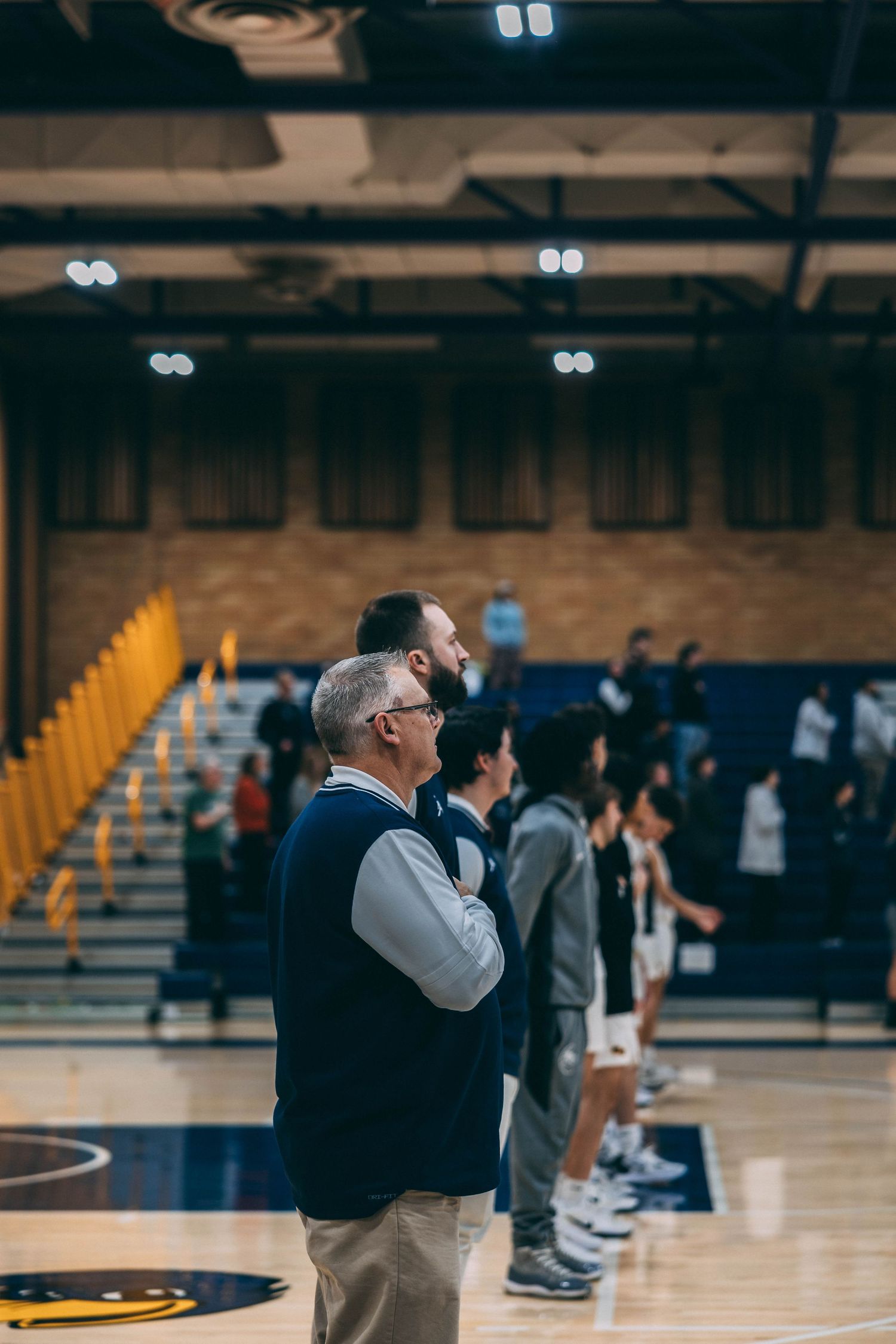 Coach observes basketball practice from sideline in gymnasium with yellow bleachers.
